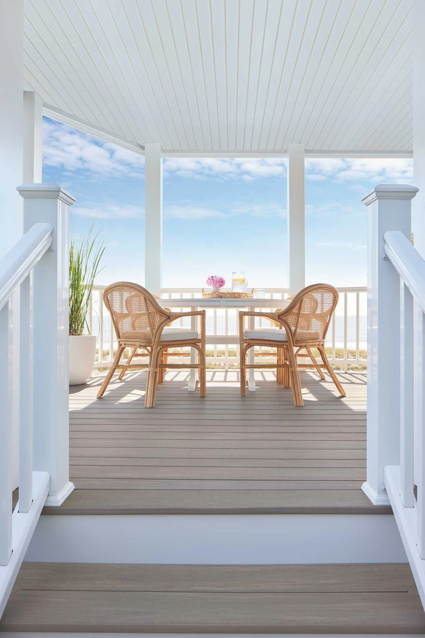 A cozy balcony with two wicker chairs and a small table set with a flower bouquet, a pitcher, and glasses, overlooking a blue sky with some clouds.