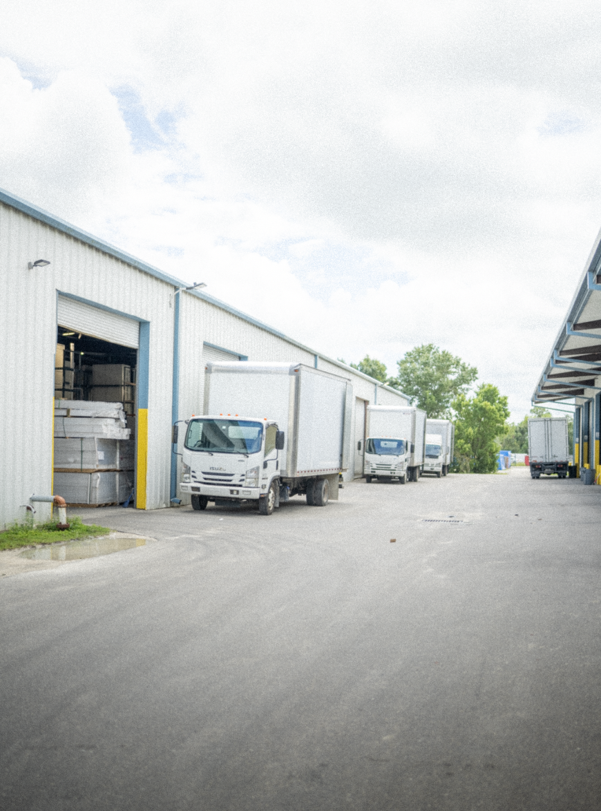 Commercial warehouse with delivery trucks parked outside, some unloading goods, overcast sky.