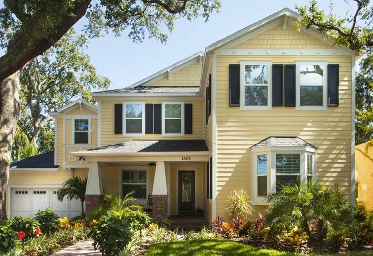 Yellow two-story house with black shutters, a gabled roof, and a front porch, surrounded by a landscaped garden with plants and flowers.