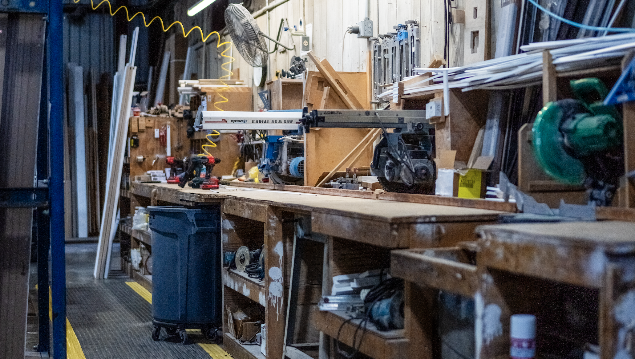 A cluttered woodworking workshop with various power tools, wood planks, and equipment on a wooden workbench.