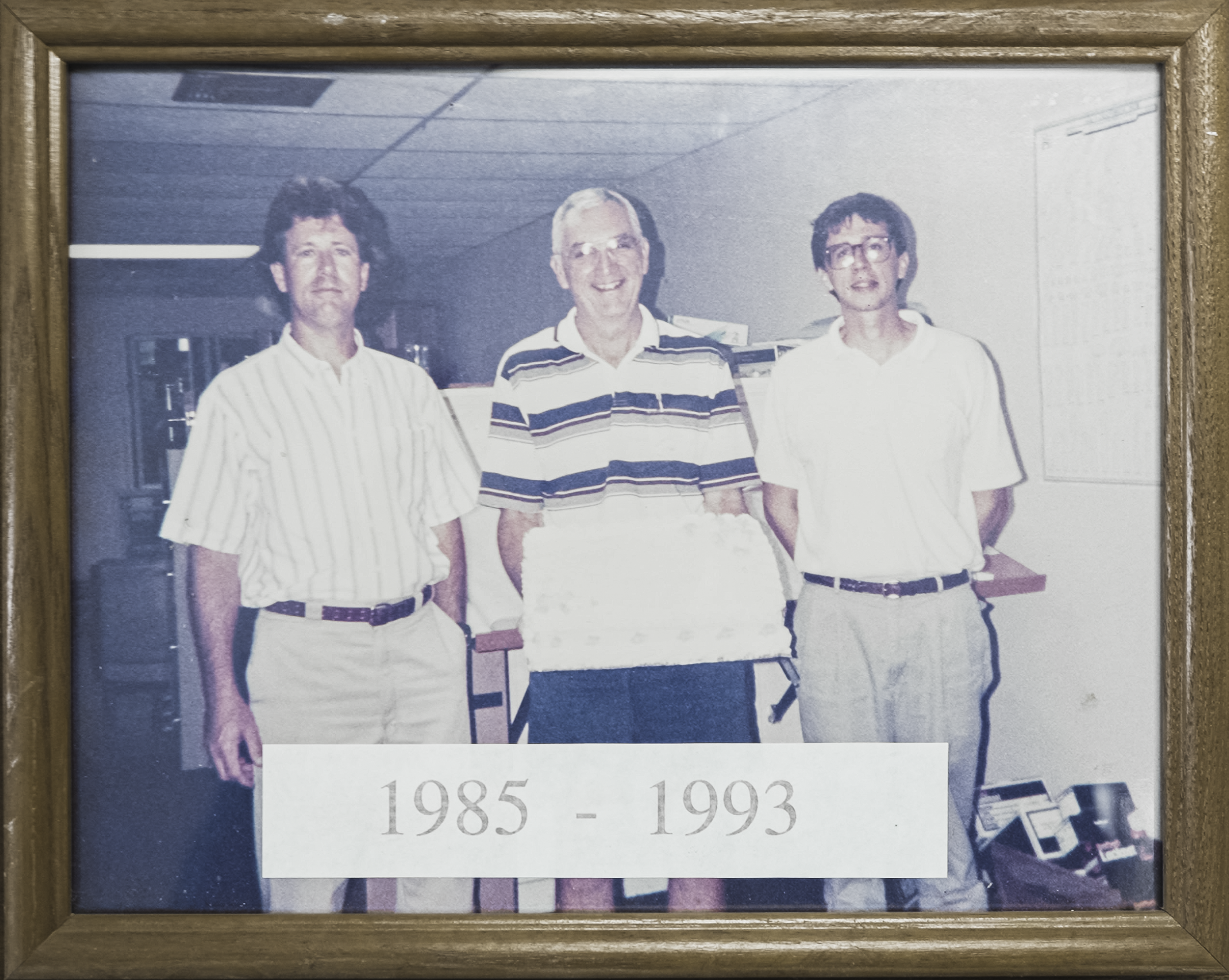 Photograph of three men standing indoors, with the years 1985-1993 printed at the bottom.