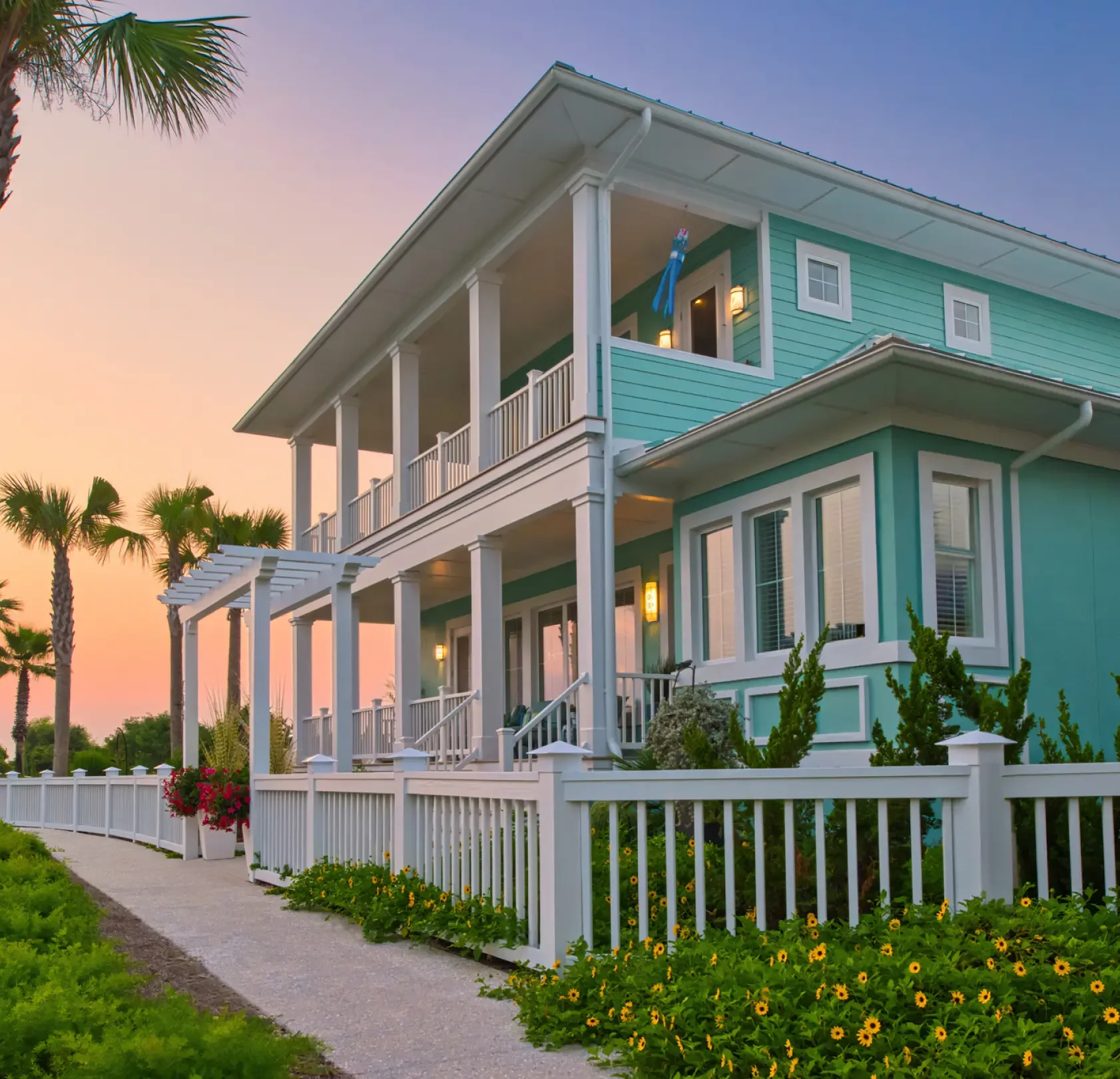 A two-story turquoise house with white trim, white columns, and a white picket fence, set against a sunset sky with palm trees in the background.