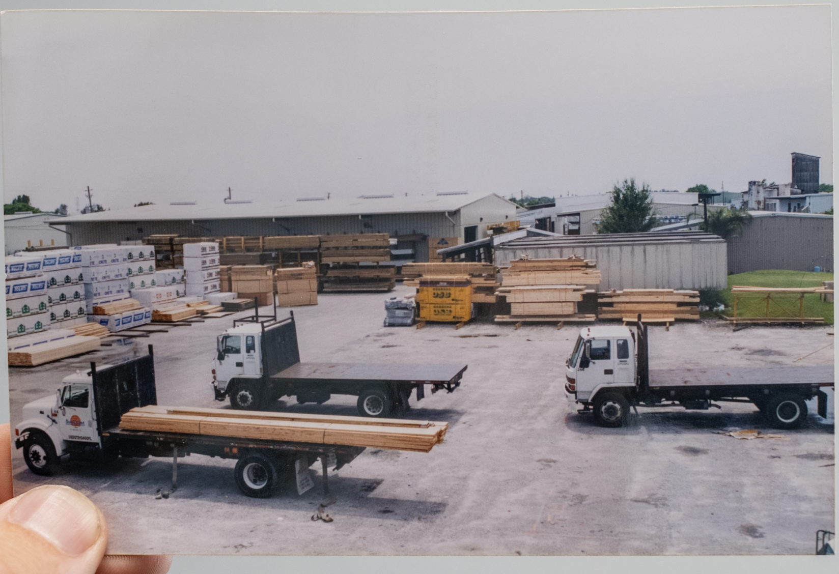 Construction yard with trucks carrying lumber, stacks of wood and building materials, metal sheets, and multiple warehouse buildings in the background, under a mostly cloudy sky.