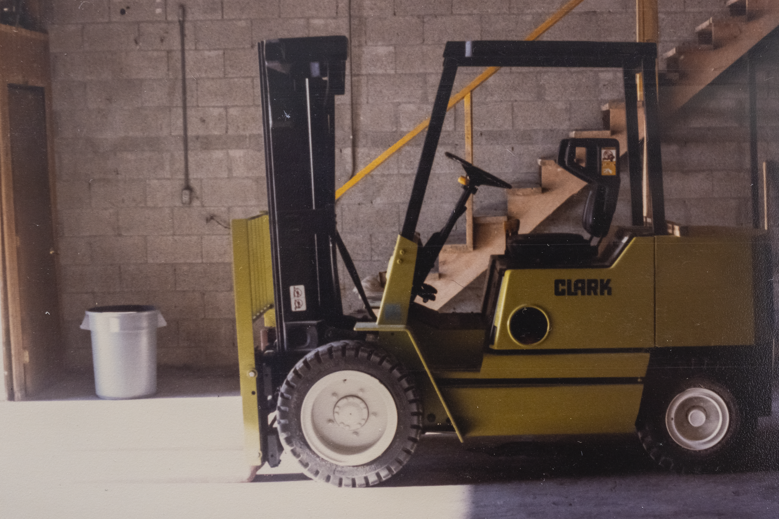 A yellow Clark forklift with black accents parked indoors against a brick wall. The forklift has a small operator seat and a steering wheel, and is positioned beside a staircase. A metal trash can is on the left side of the image.