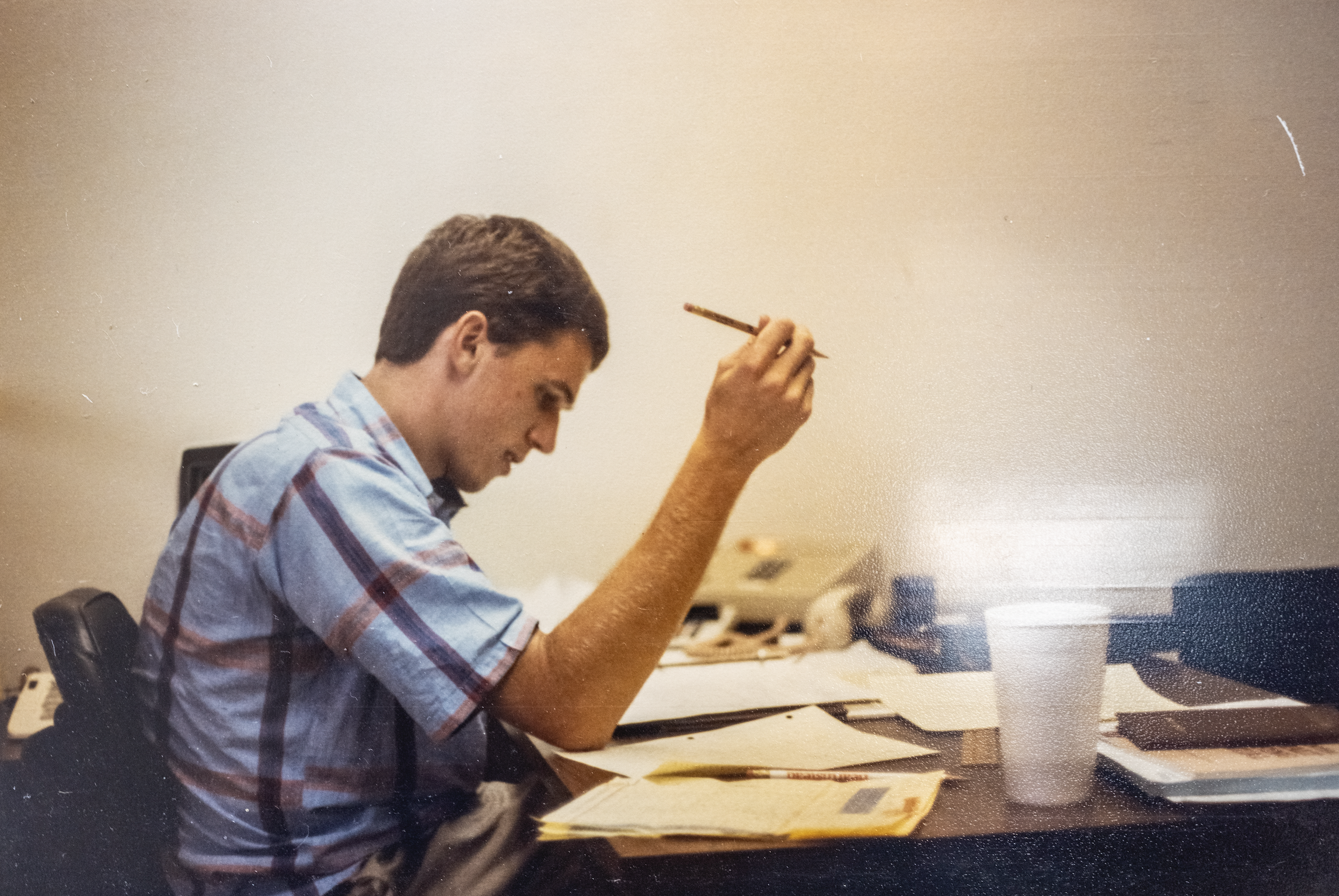 A young man with short brown hair, wearing a blue plaid shirt, sitting at a cluttered desk with papers, notebooks, a white cup, and other office supplies, reading a document and holding a pen.