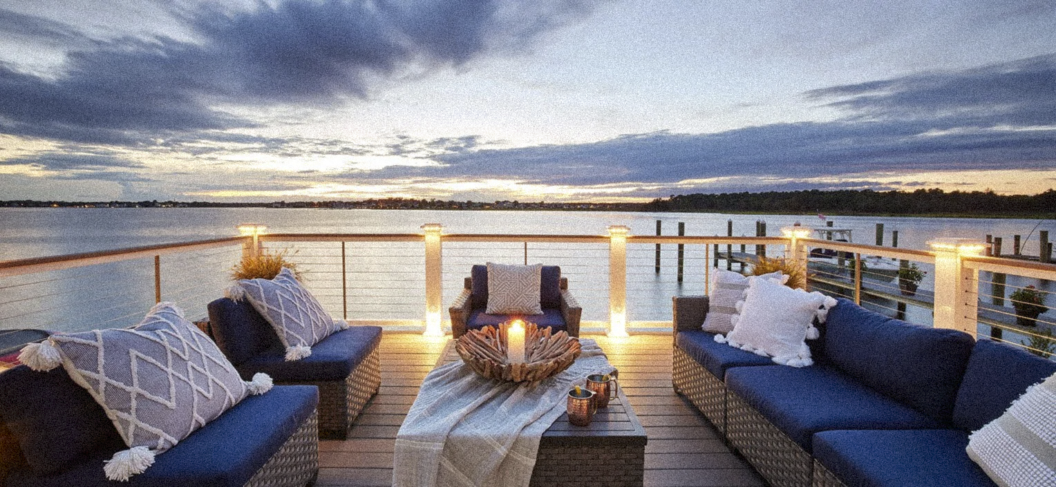 Outdoor patio overlooking a river at sunset with wicker furniture, blue cushions, pillows, a candle, and string lights.