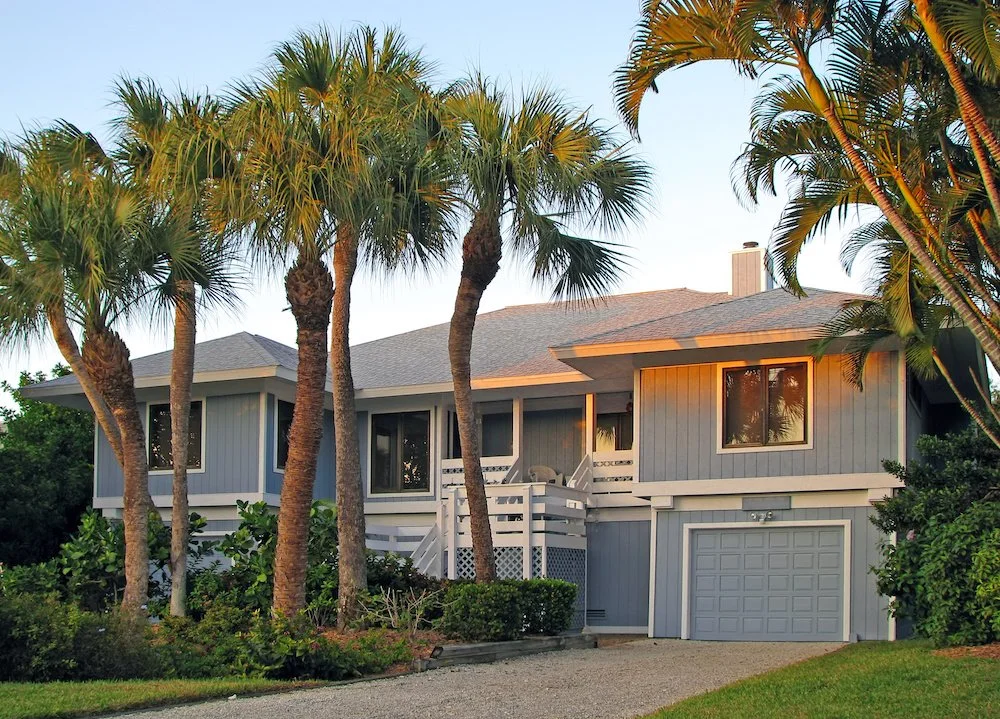 A two-story house painted in light blue with palm trees in front, a sloped roof, and a one-car garage, with a well-maintained lawn and landscaping.