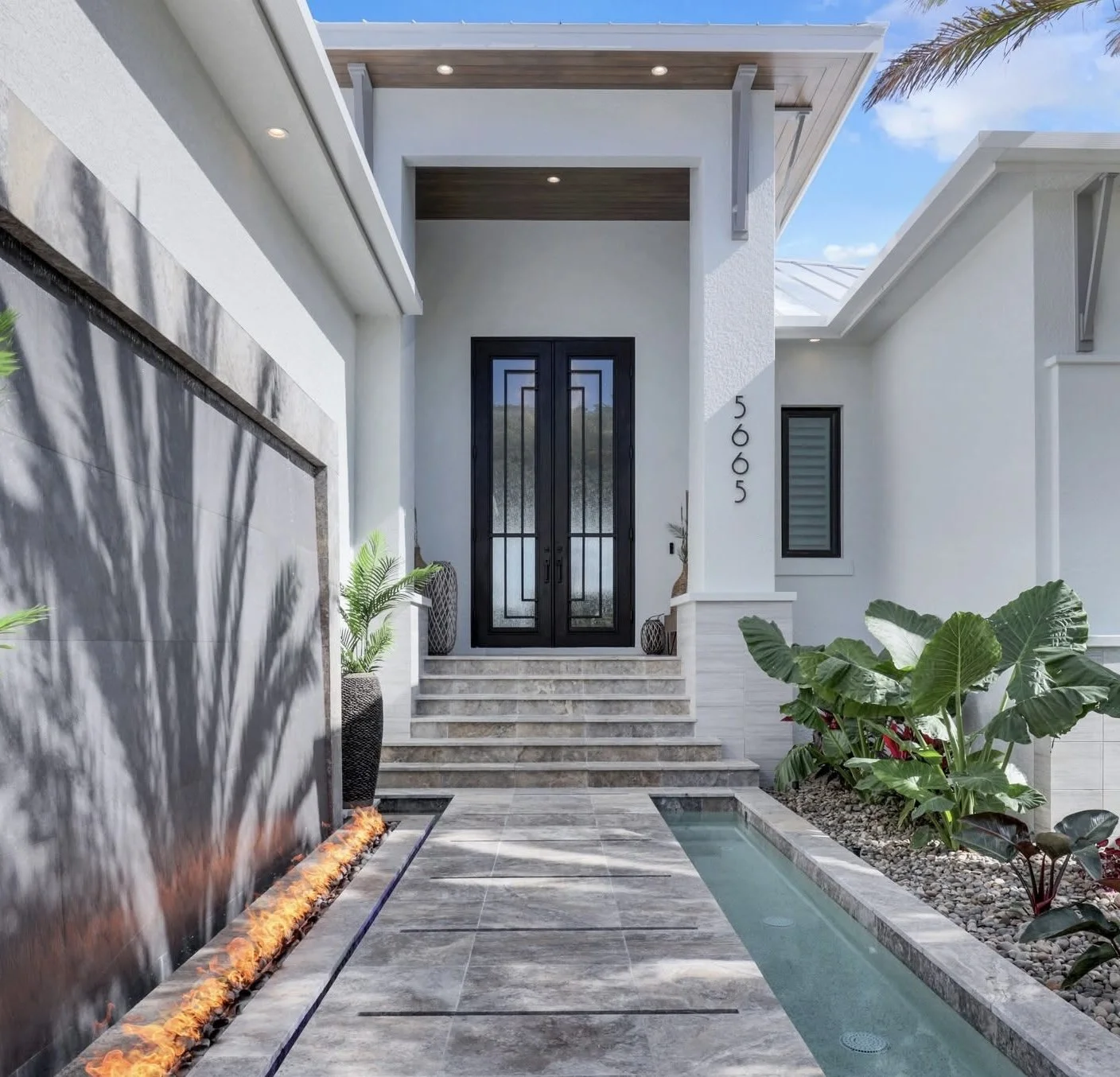 Modern house entrance with black double doors, white exterior walls, steps leading to the door, and a small water feature and tropical plants in the front yard.