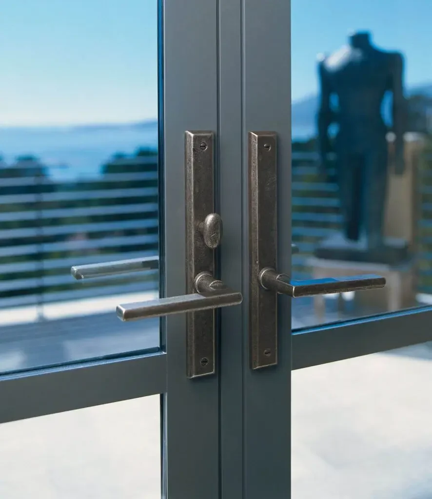 Close-up of glass door handles with a view of a balcony and ocean outside, with a blurred figure in black clothing standing on the balcony.