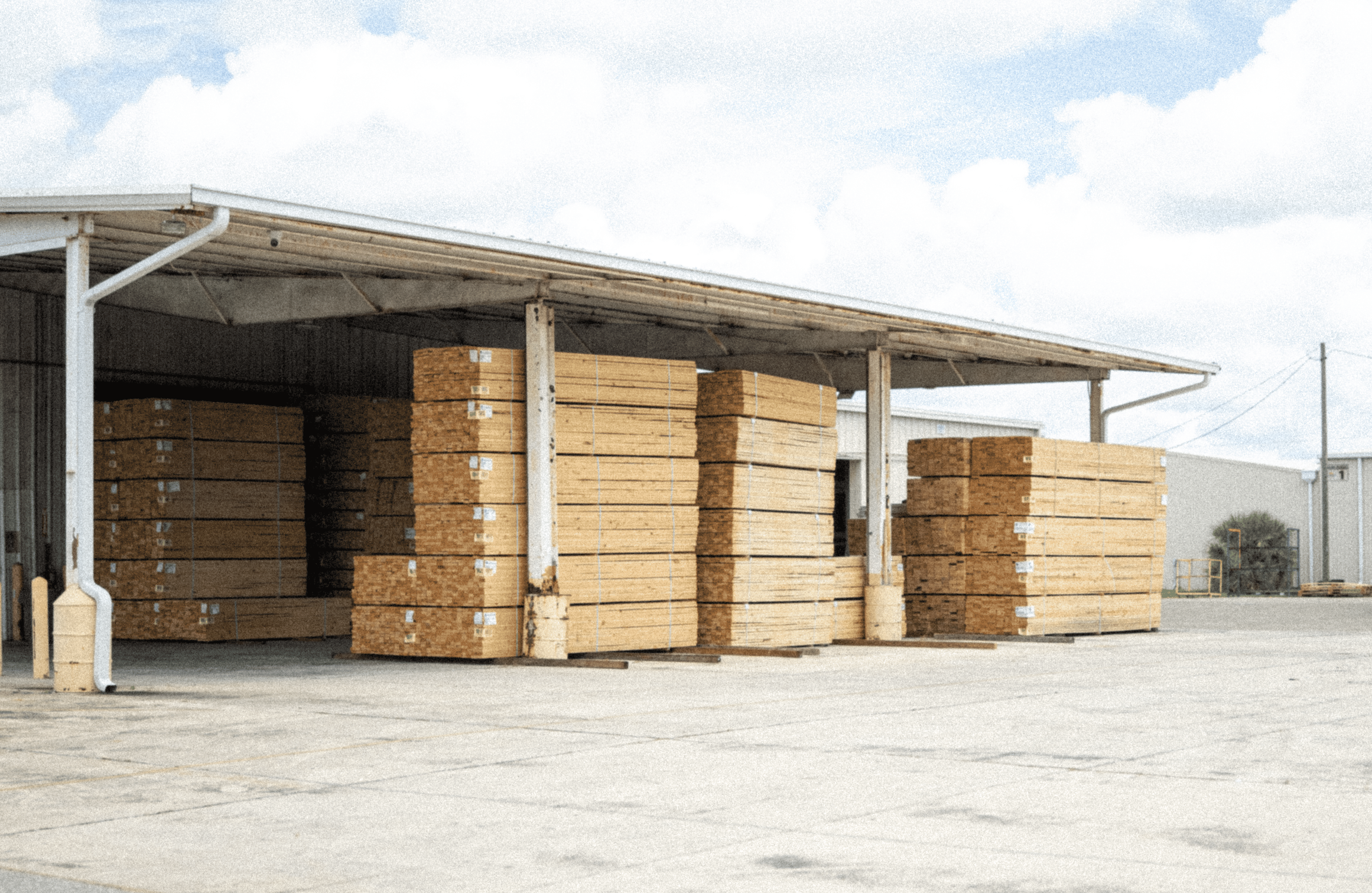 Stacks of lumber stored under a metal shed in an outdoor storage yard with a cloudy sky in the background.