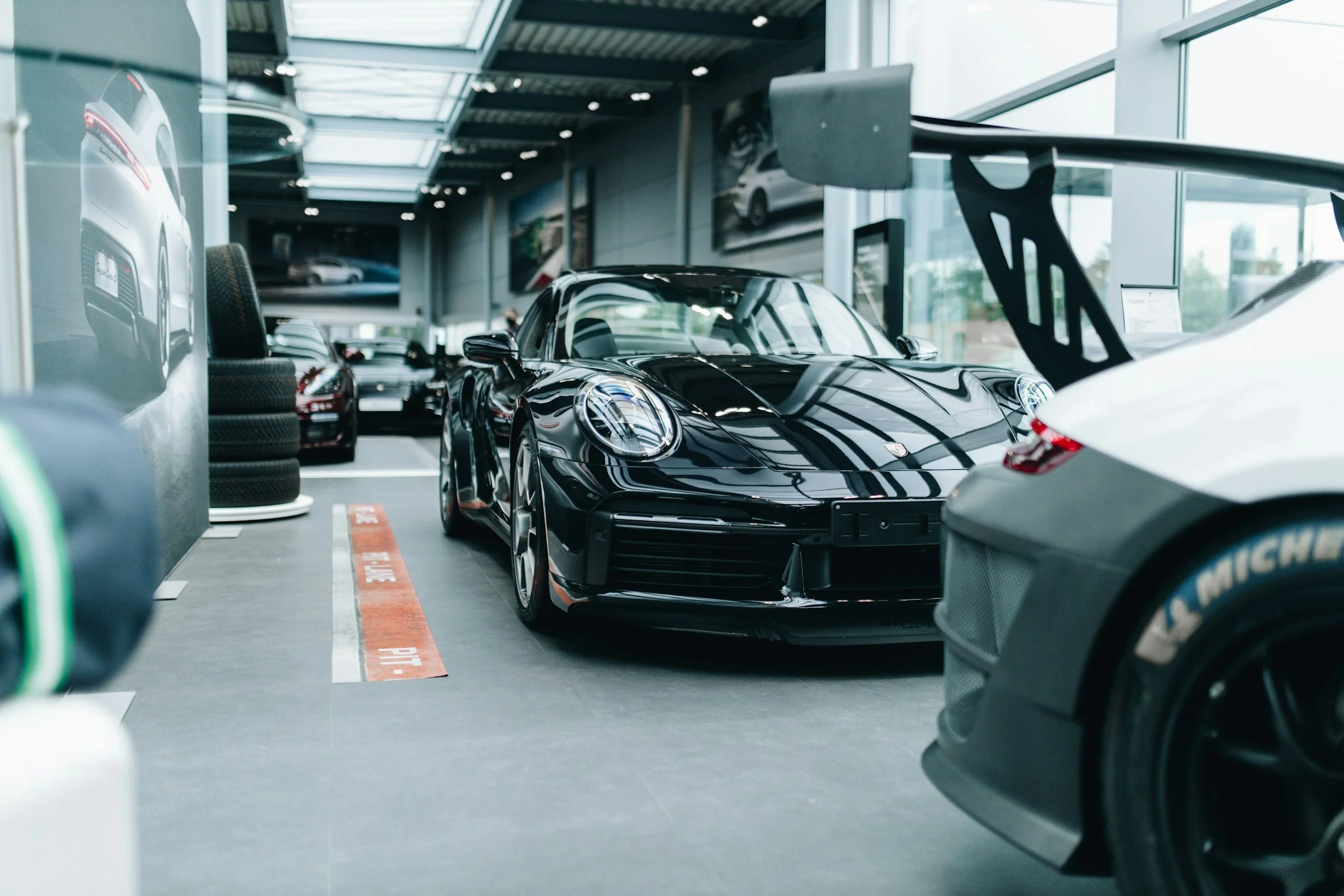 Interior of a modern car showroom with several cars on display, including a black sports car in the centre, large windows letting in natural light, and various automotive branding and signage.