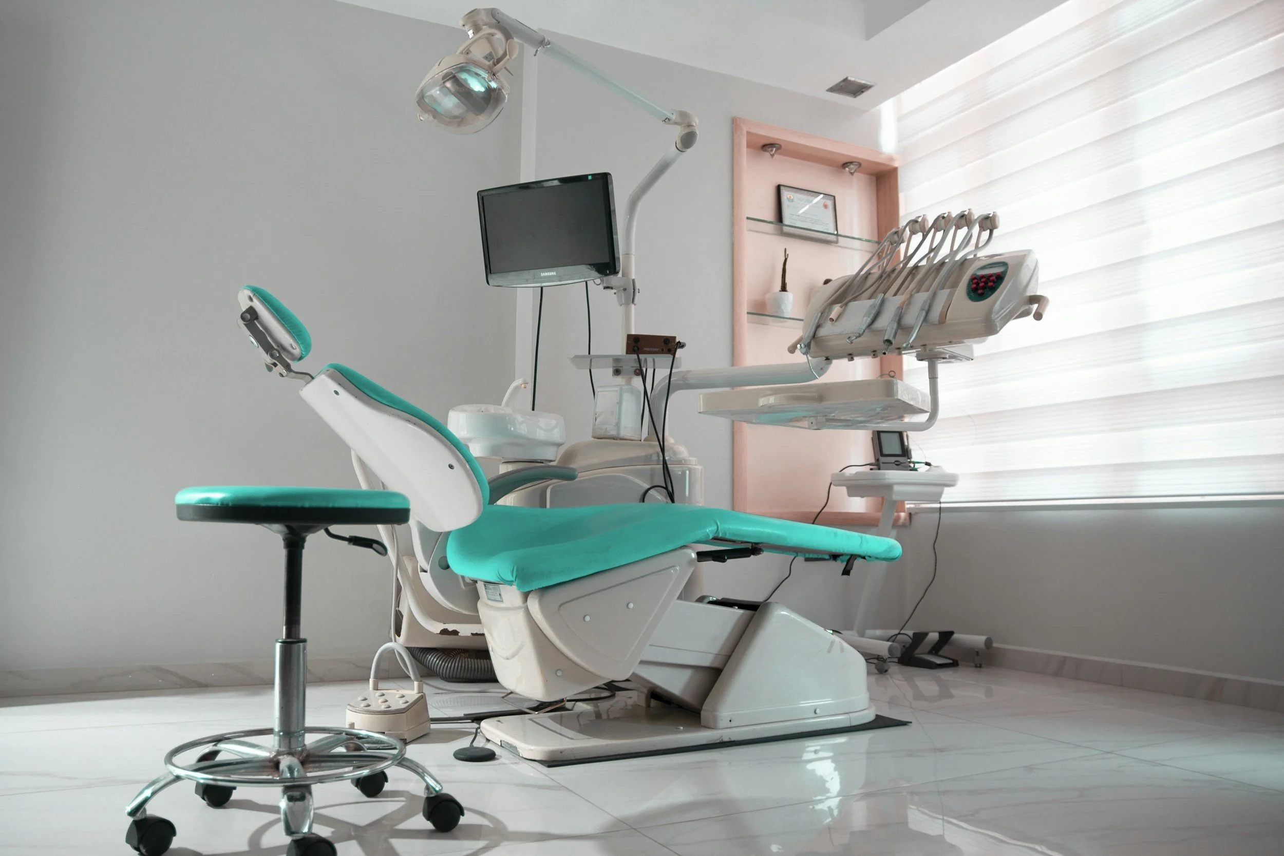 An empty dental clinic room showing a dental chair with teal cushioning, a stool, and dental equipment including a monitor, light, and tools, with window blinds in the background.