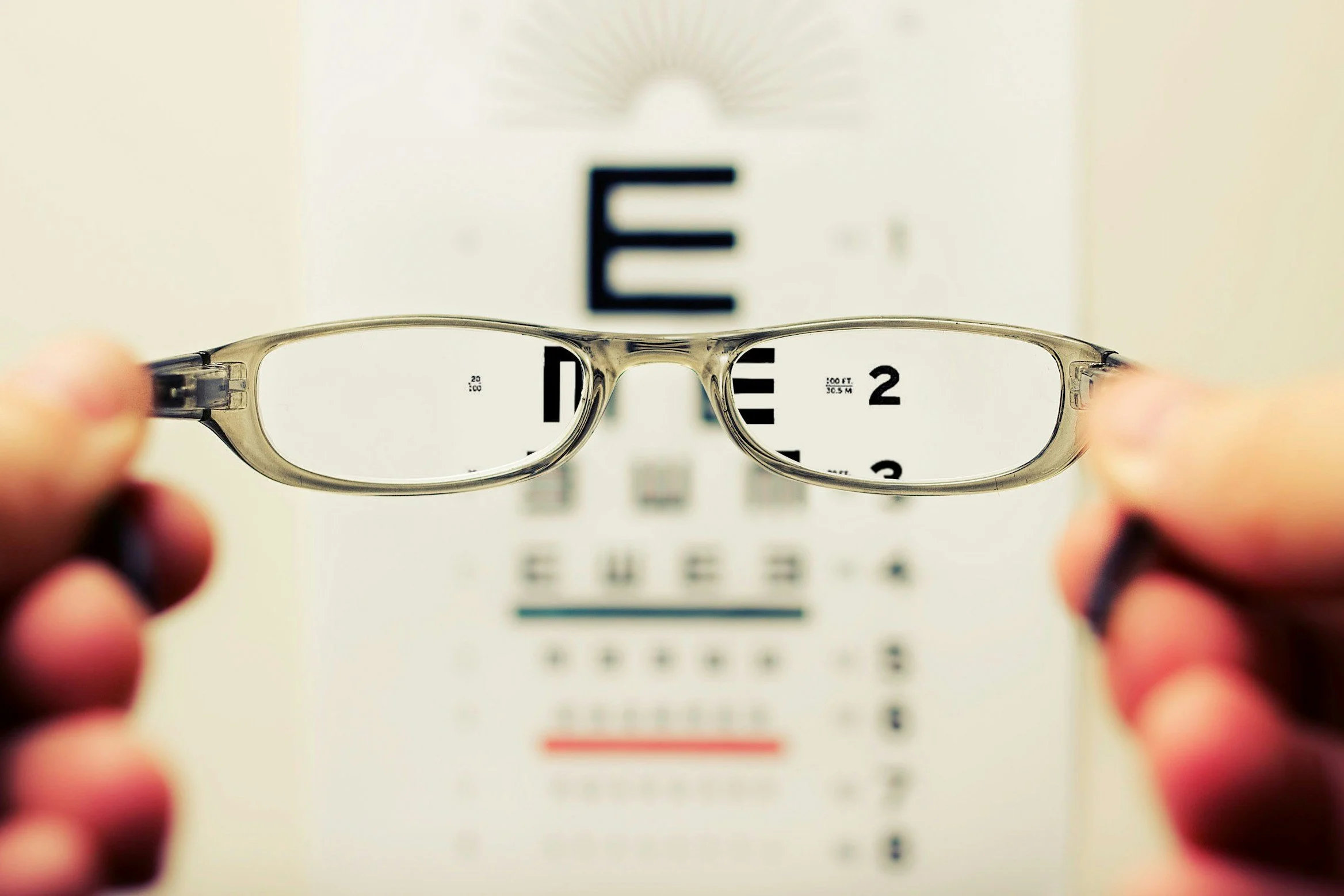 A pair of eyeglasses held in front of an eye chart used for eyesight testing, with blurred letters visible through the glasses.