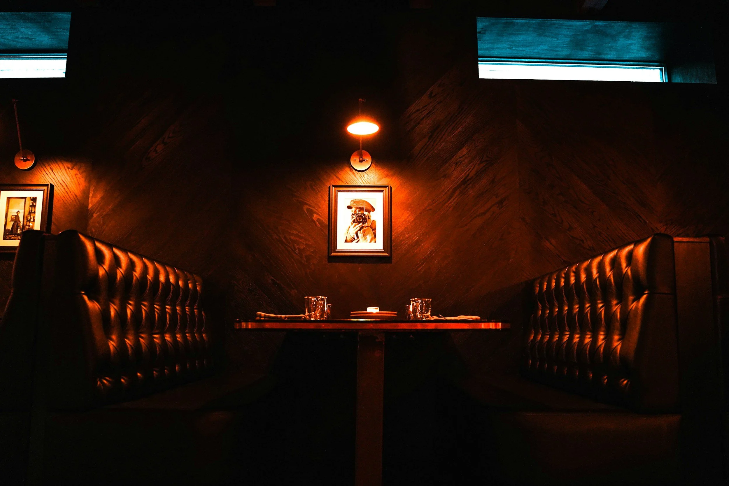 A cozy restaurant booth with tufted leather seating, a wooden table with glasses and utensils, dark wood-paneled walls, framed art, and warm overhead lighting with two windows at the top.