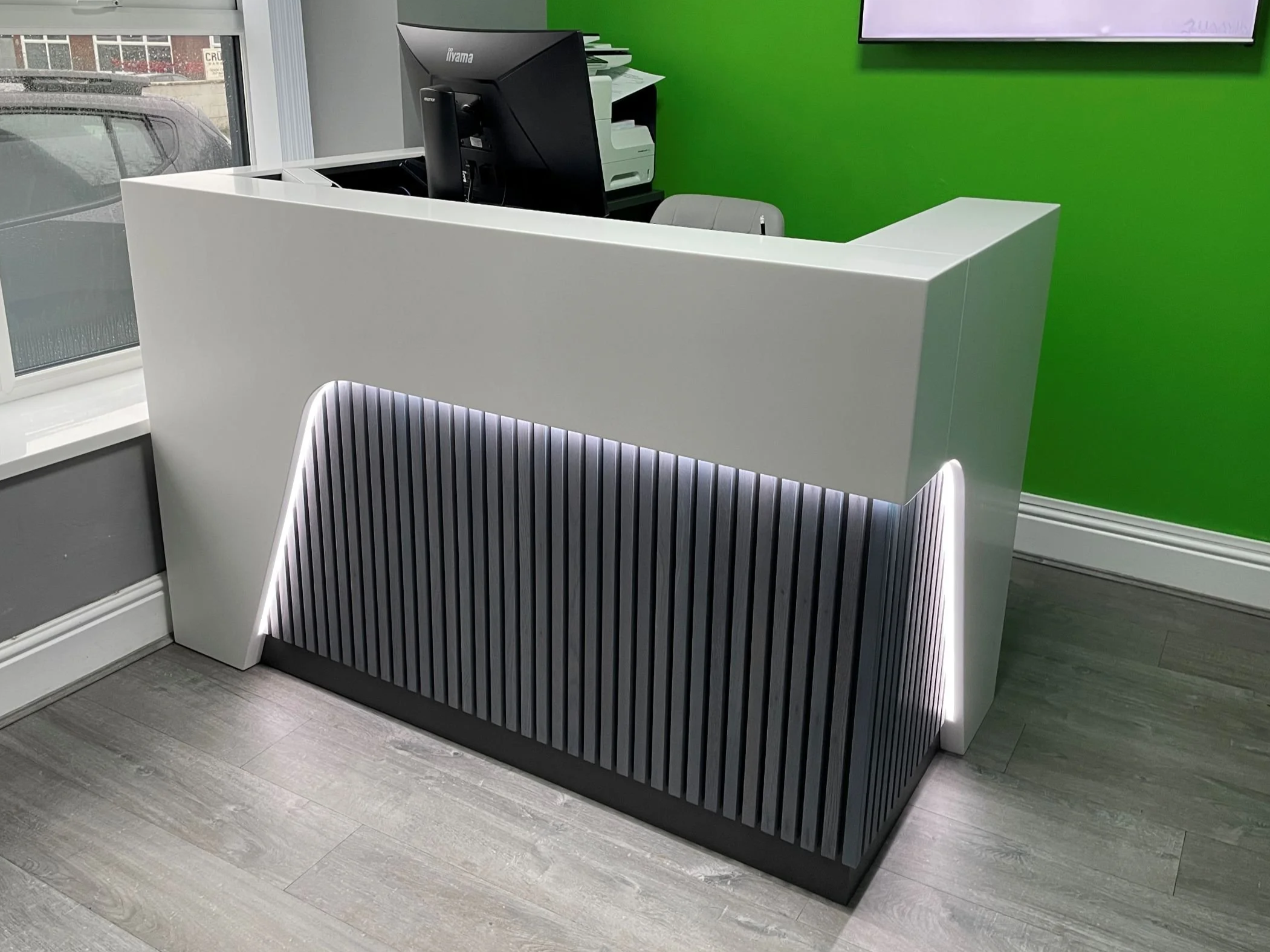 Modern bespoke reception desk with dark grey stained Oak vertical slats to front panel and solid surface finishes to upper sections. Installed in the reception area of SpineFine clinic, room has green walls and light grey woodgrain flooring.