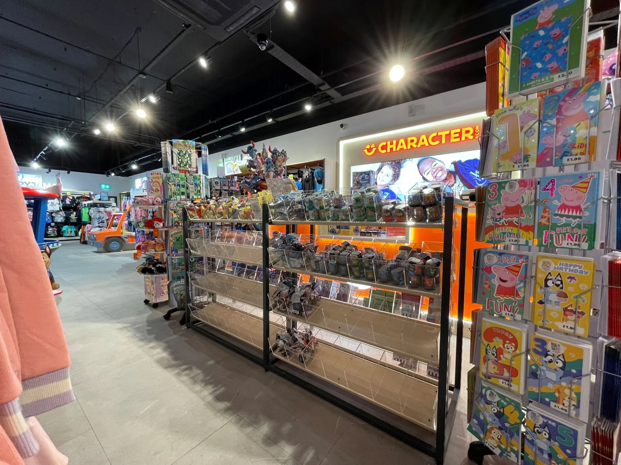 Character.com retail store section with an LED illuminted sign reading 'CHARACTER.COM', shelves filled with toys and games, colourful gift bags, and a display of Peppa Pig birthday cards. Bespoke metal retail display fittings in foreground.