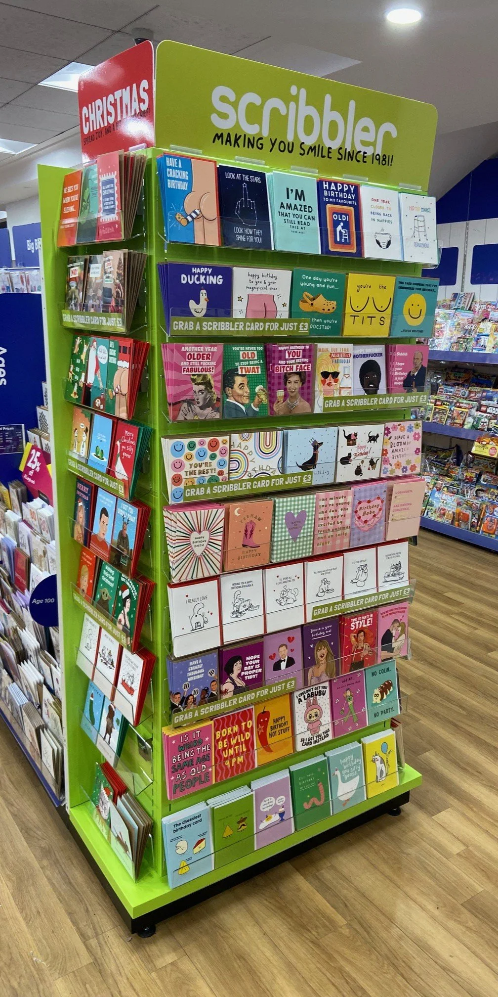 A colourful bespoke metal retail concession display stand displaying Scribbler cards in a host store. The stand is green with designated sections for different types of cards, and a sign at the top reads 'scribbler making you smile since 1981!'.