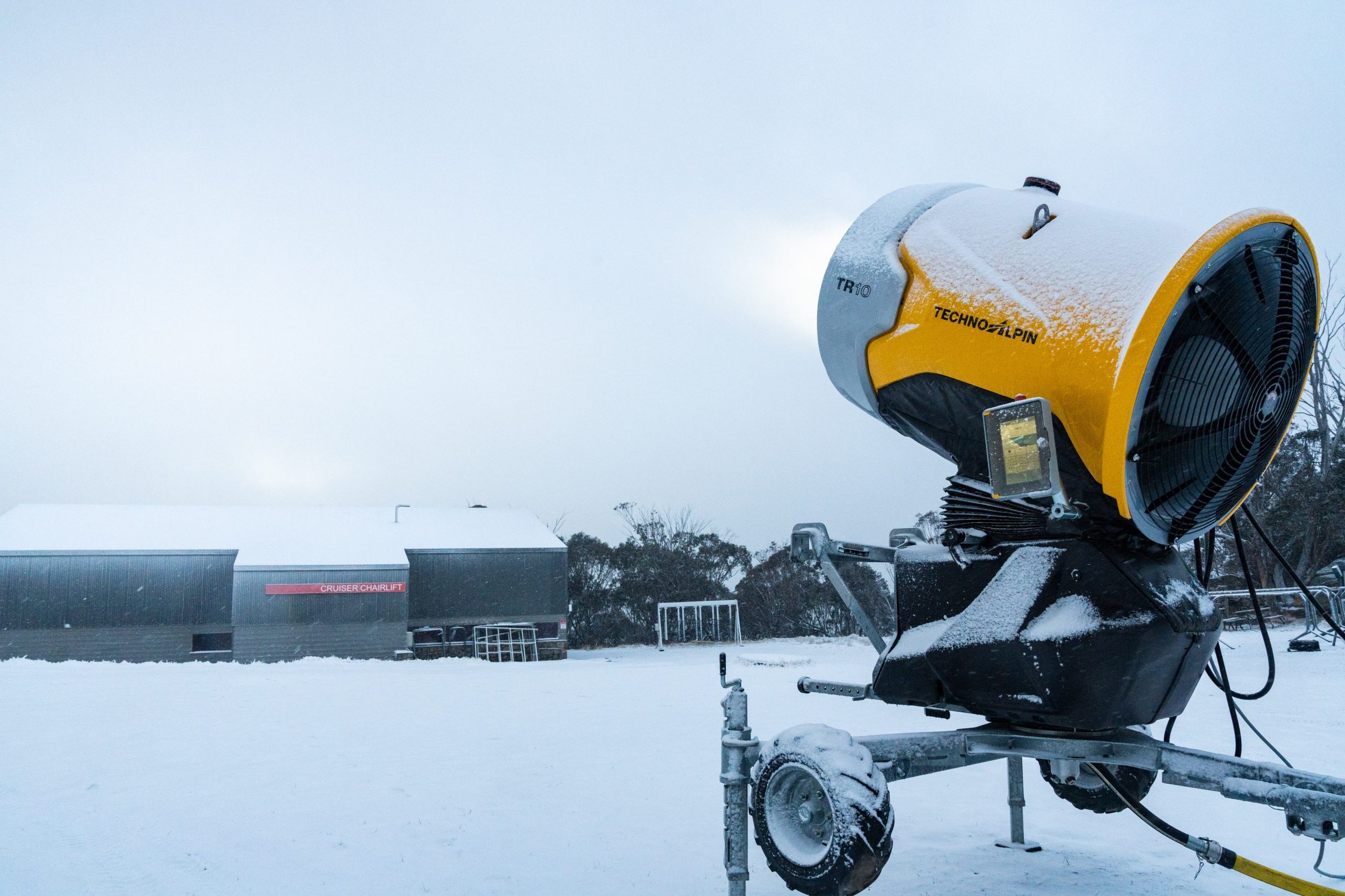 Yellow and black industrial fan with snow on it, outdoors in a snowy area with a building in the background.