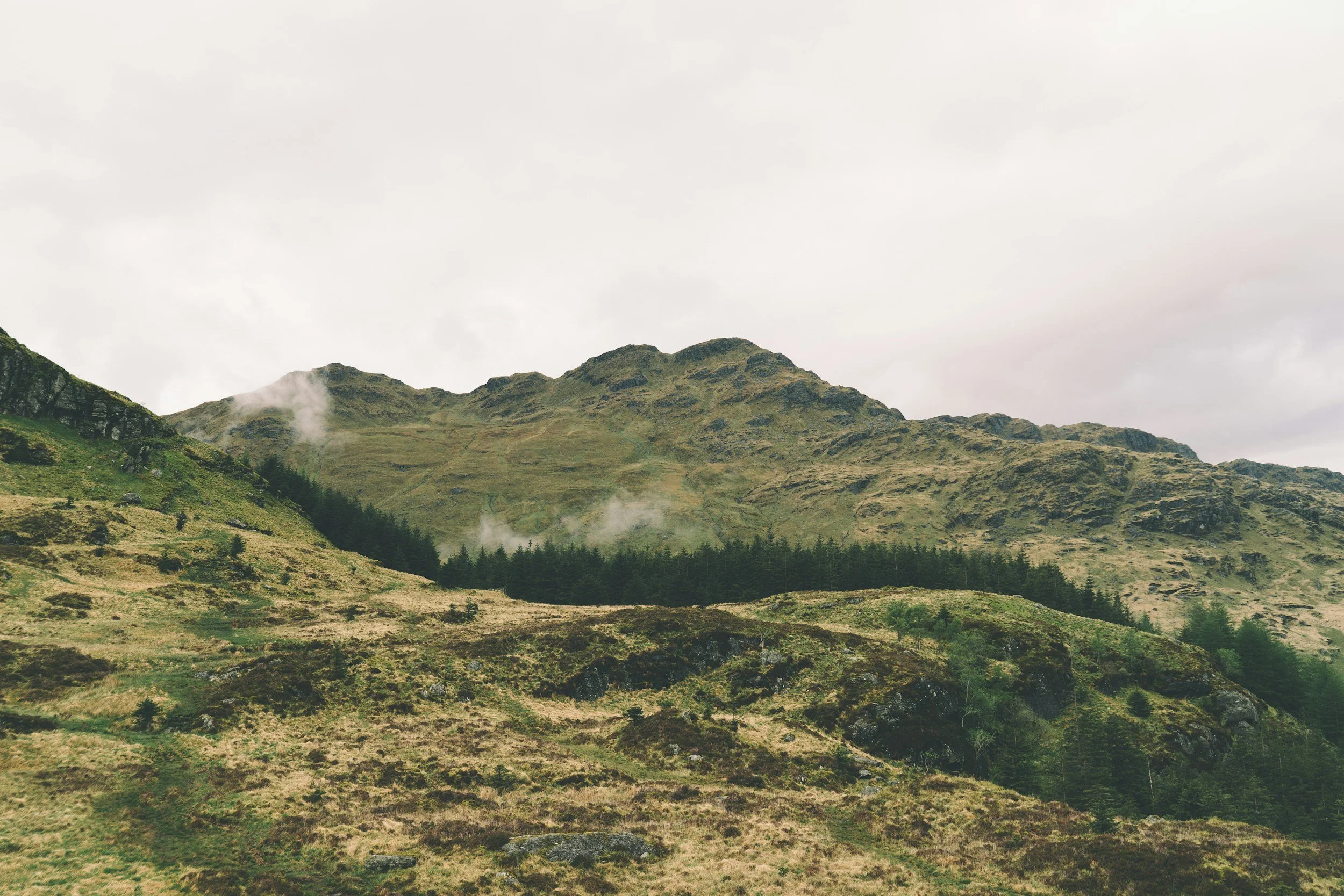 A green mountainous landscape with hills and rocky outcroppings, some trees, under a cloudy sky.