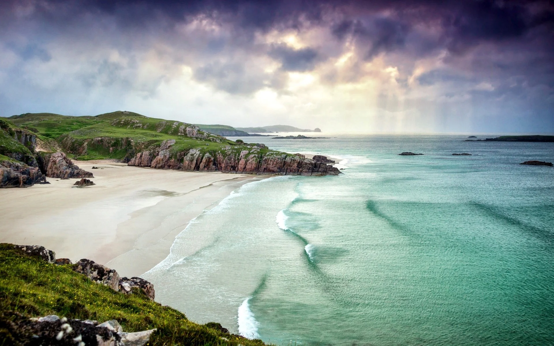 Scenic view of a beach with white sand, green grassy cliffs, rocks, ocean waves, and a cloudy sky over the horizon.