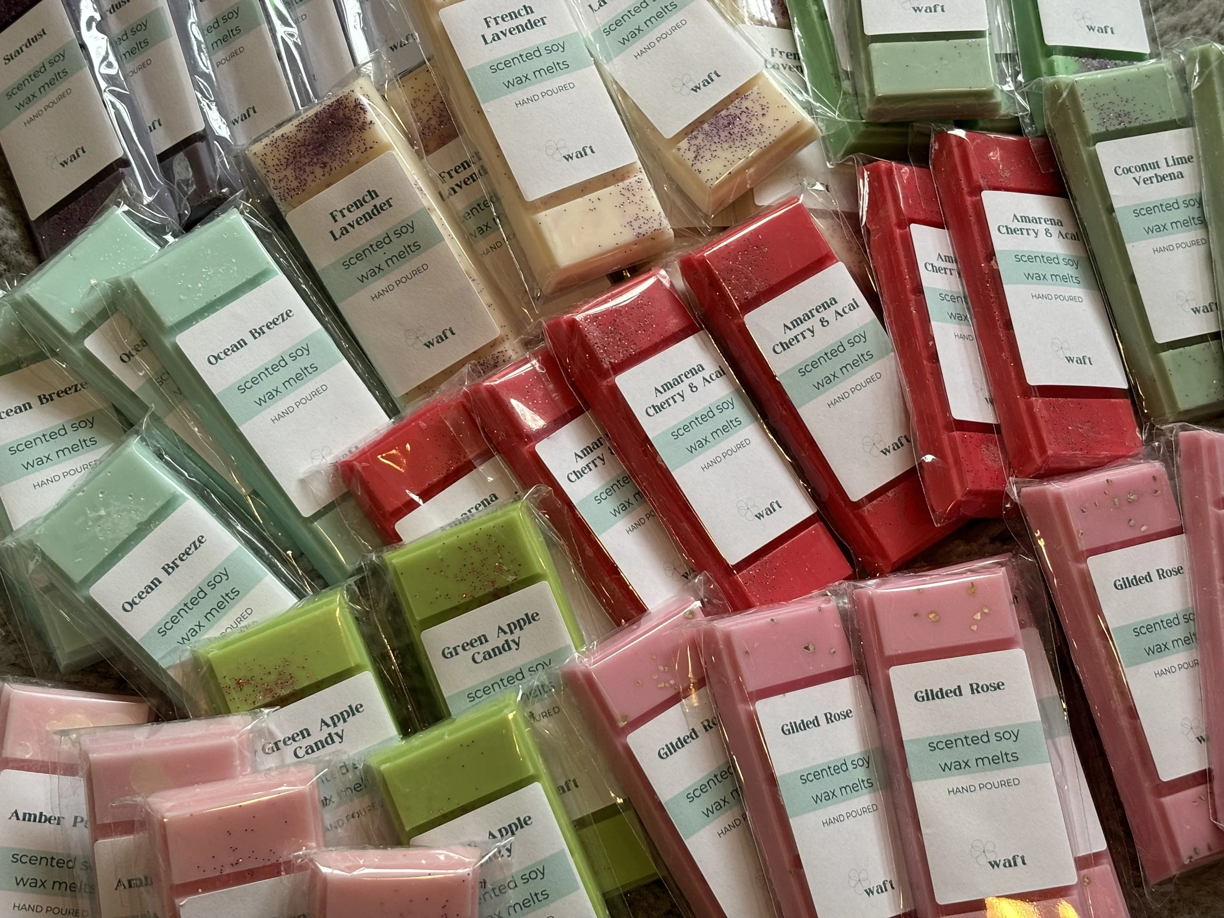 A collection of colorful scented soy wax melts in various flavors including Ocean Breeze, Green Apple Candy, Gilded Rose, Amarena Cherry & Açaí, French Lavender, and Coconut Lime Verbena, all individually wrapped in clear plastic with labels.