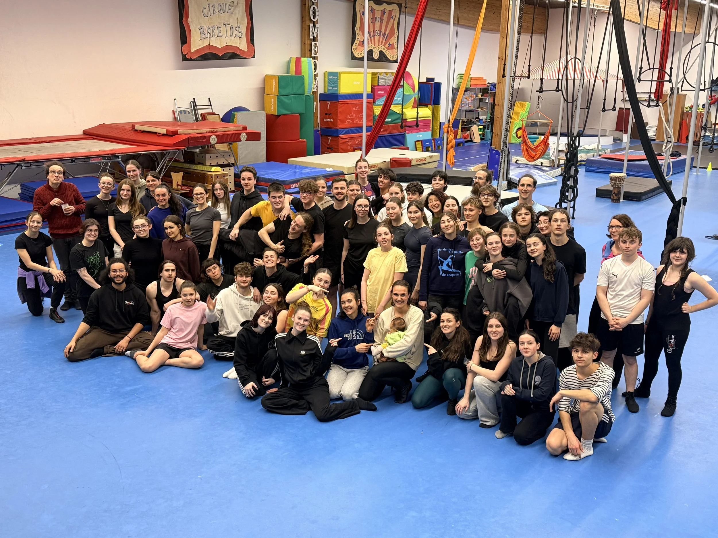 Un groupe de personnes, principalement jeunes, rassemblées dans une salle de gymnastique ou de cirque, avec des équipements de gymnastique en arrière-plan, posent pour une photo de groupe. Certaines personnes sourient ou font des gestes amicals.