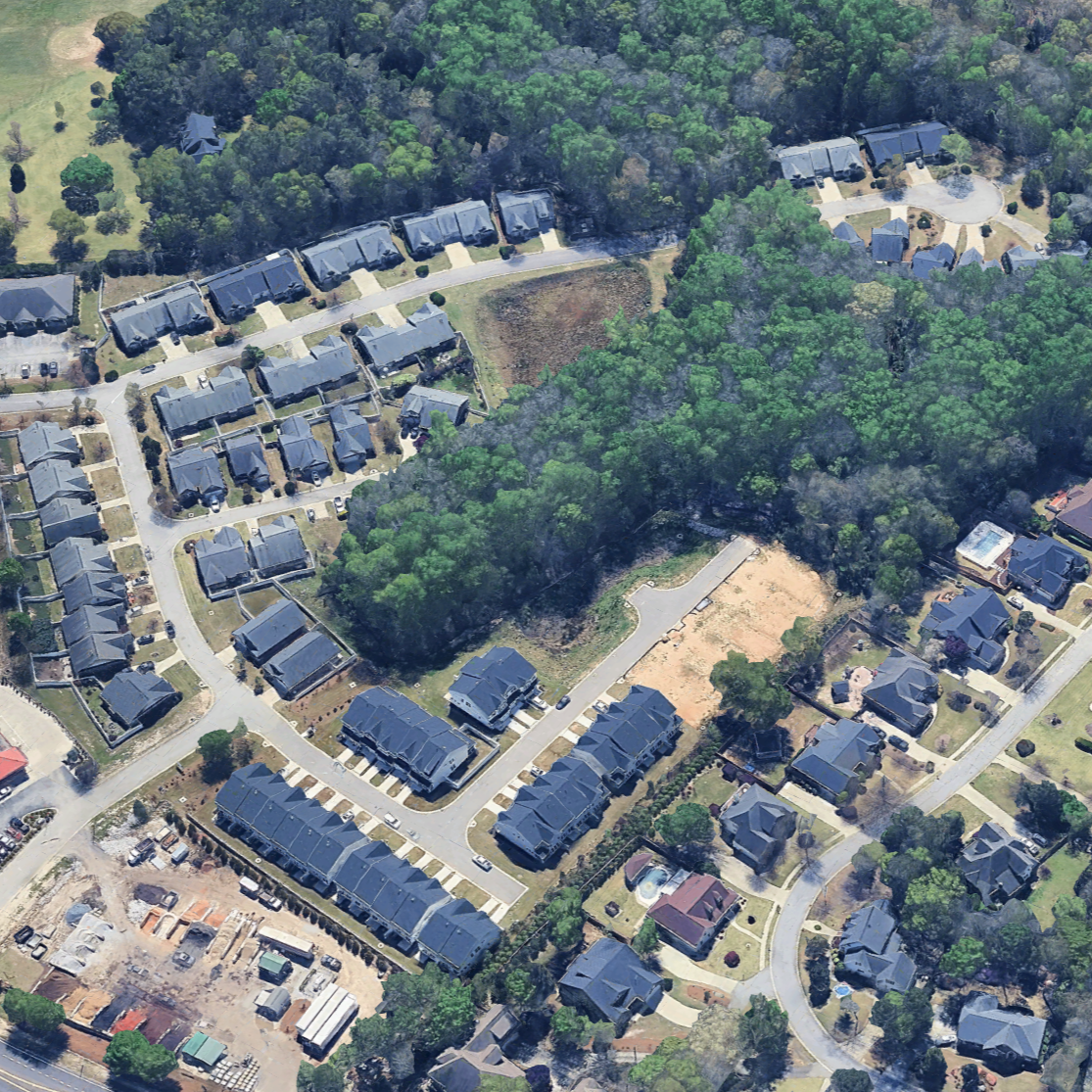 Aerial view of a suburban neighborhood with houses, streets, and green trees, including a wooded area and an empty lot.