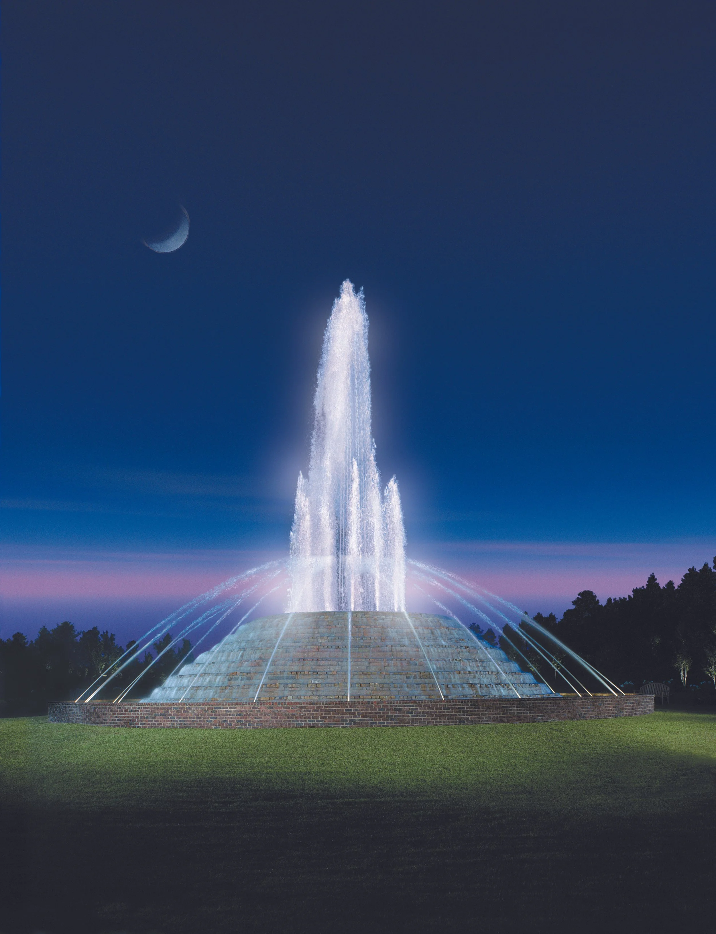 Nighttime scene of a large illuminated fountain with water jets, set on a grassy area, with a crescent moon visible in the dark sky.
