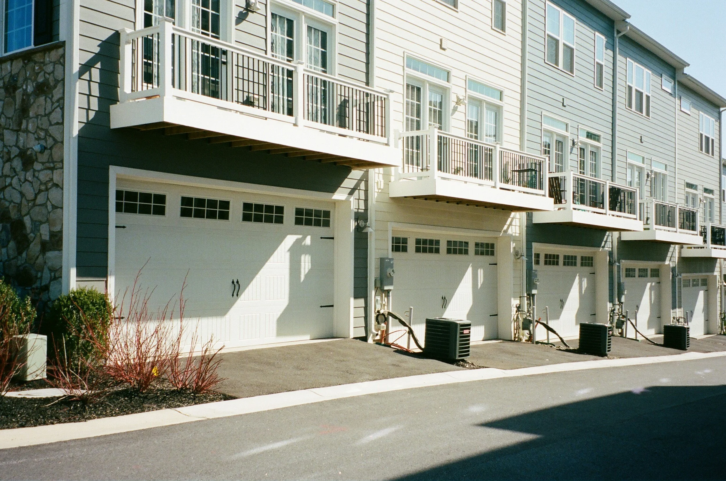 Row of modern townhouses with attached garages and small balconies, featuring stone and siding exteriors, along a paved street.