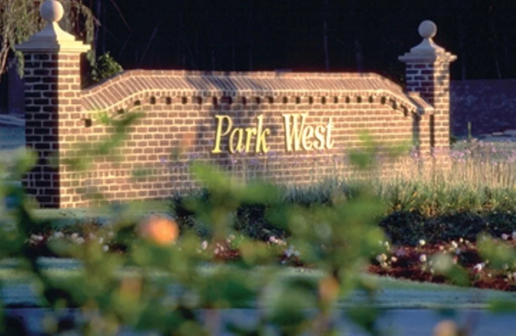 A brick entrance sign reading 'Park West' surrounded by plants and flowers, with fencing in the background.