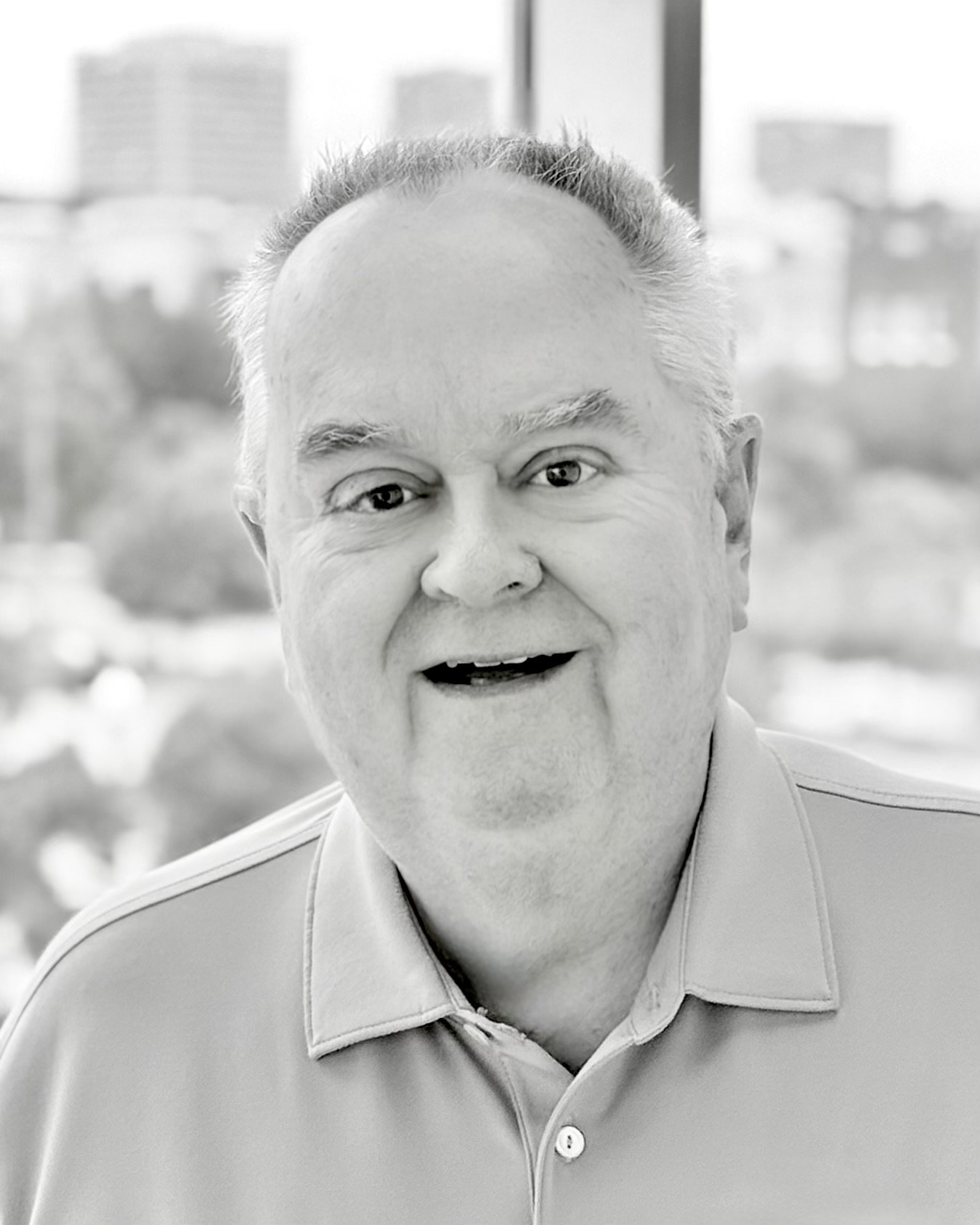 A smiling man with short hair and a collared shirt, indoors with large windows showing a cityscape in the background.