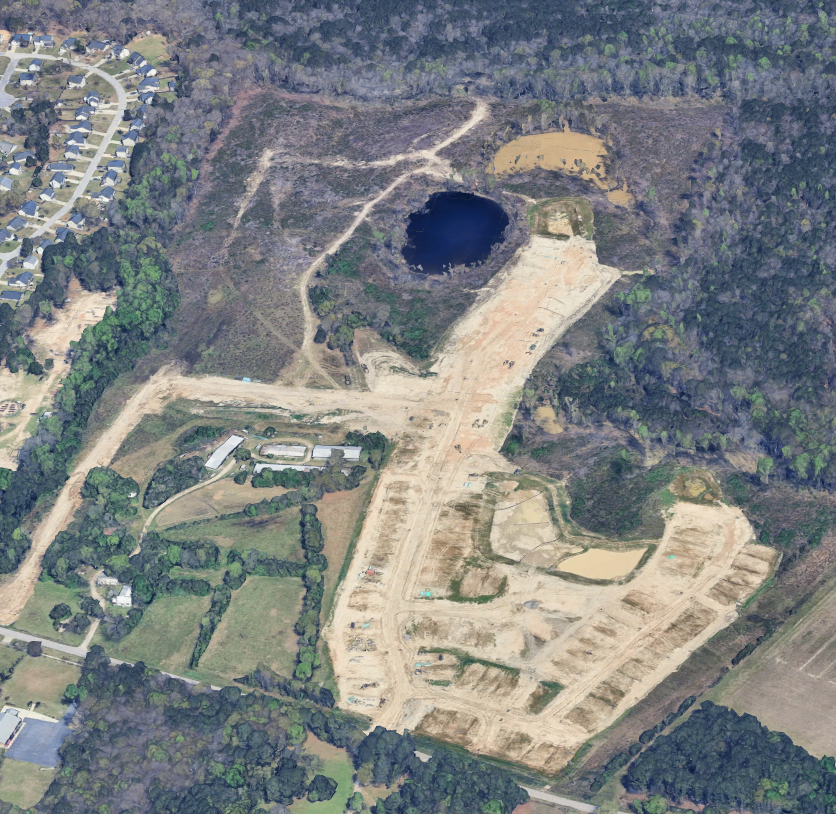 Aerial view of a construction site with cleared land, dirt roads, a small pond, and surrounding wooded areas.