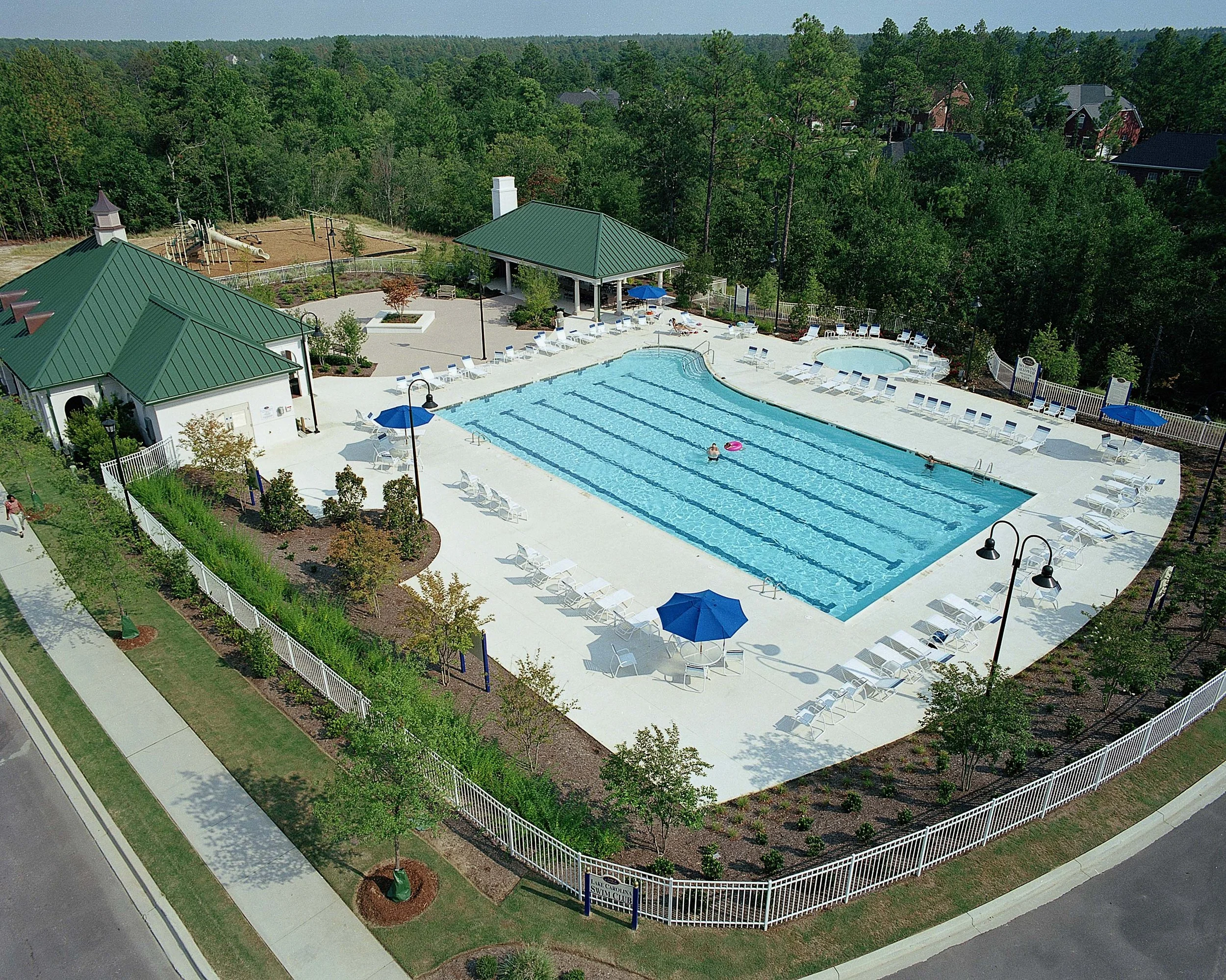 Aerial view of a community swimming pool with lounge chairs, umbrellas, and a pool house, surrounded by trees and residential houses in the background.