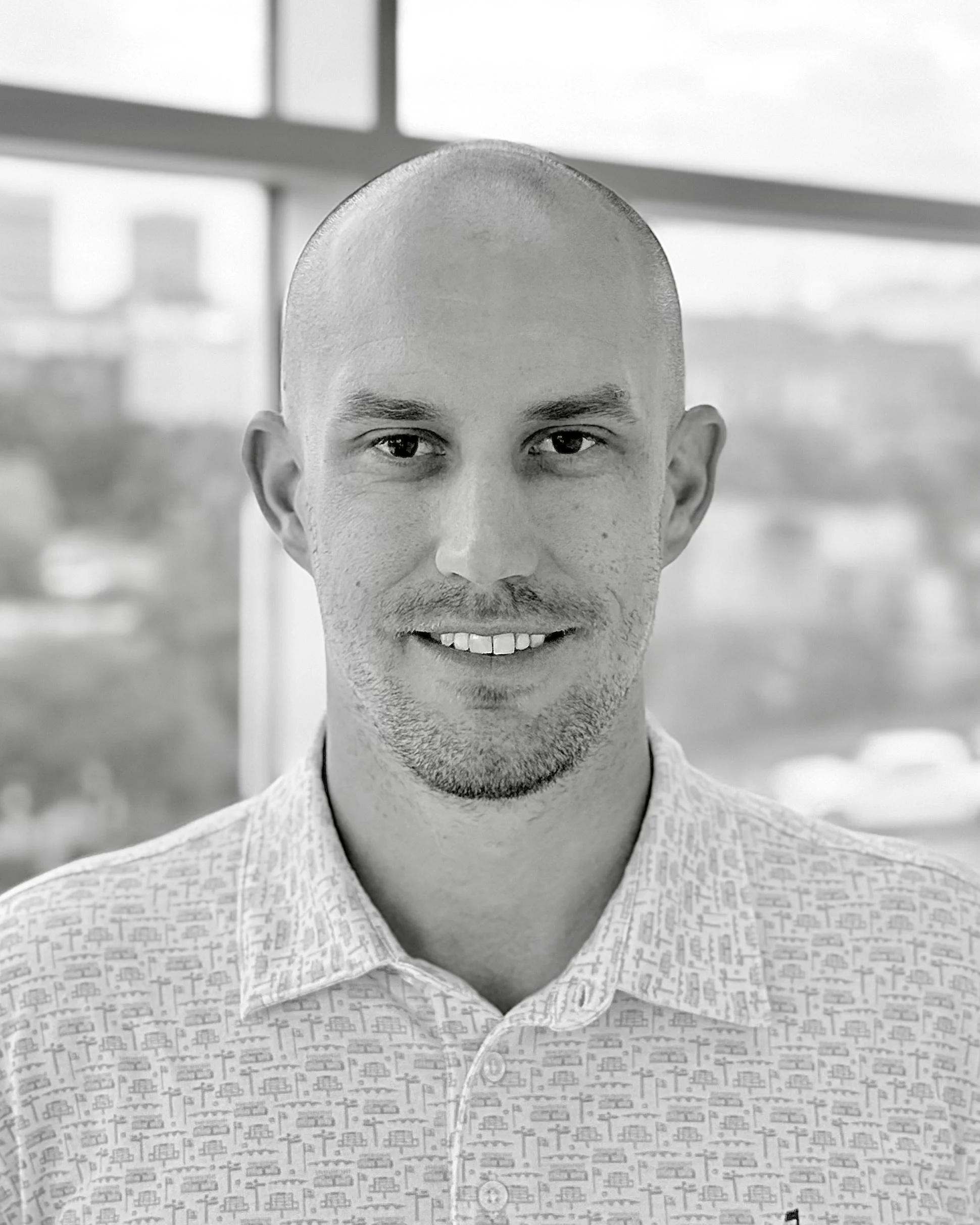 A black and white photo of a smiling man with a shaved head wearing a patterned collared shirt, standing in front of a window.