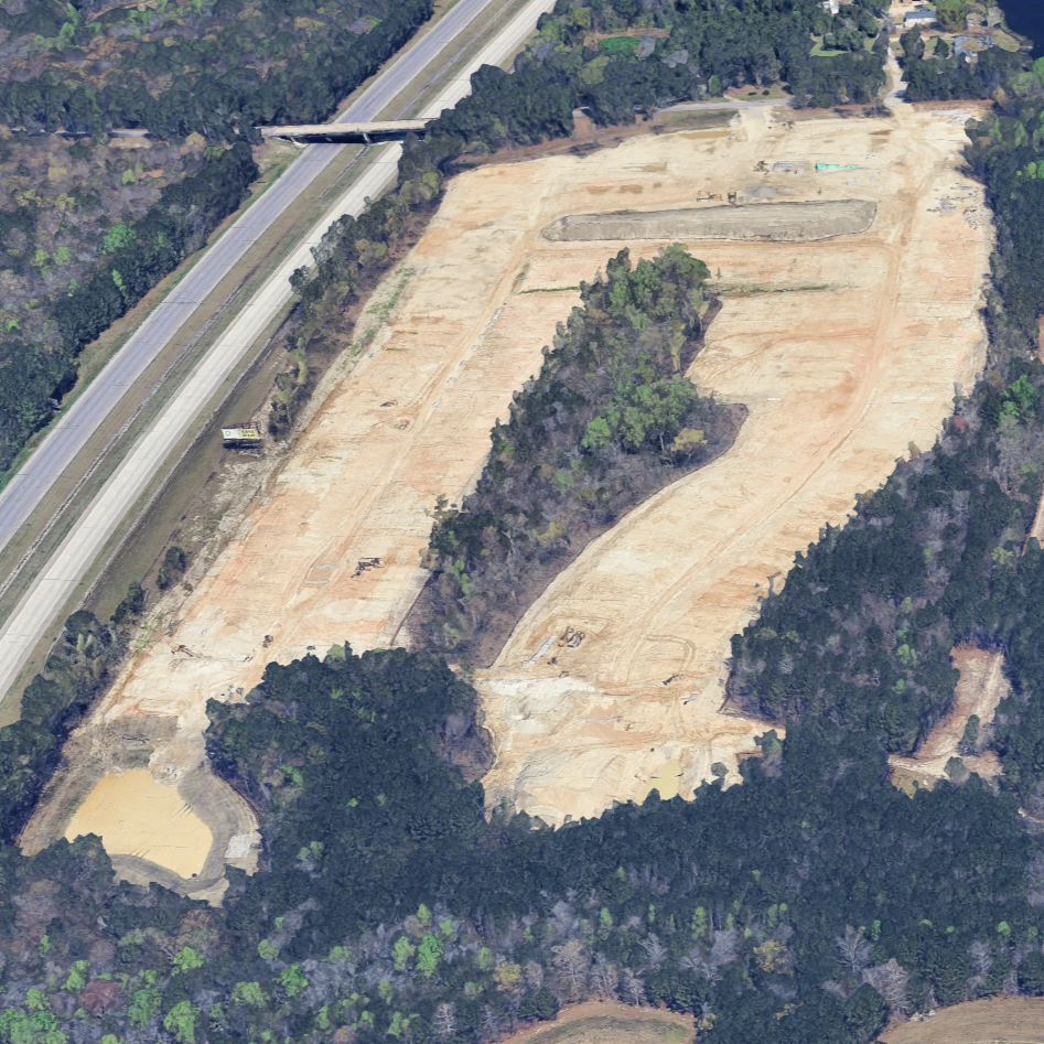 Aerial view of a construction site with cleared land, some trees, and a road overhead.