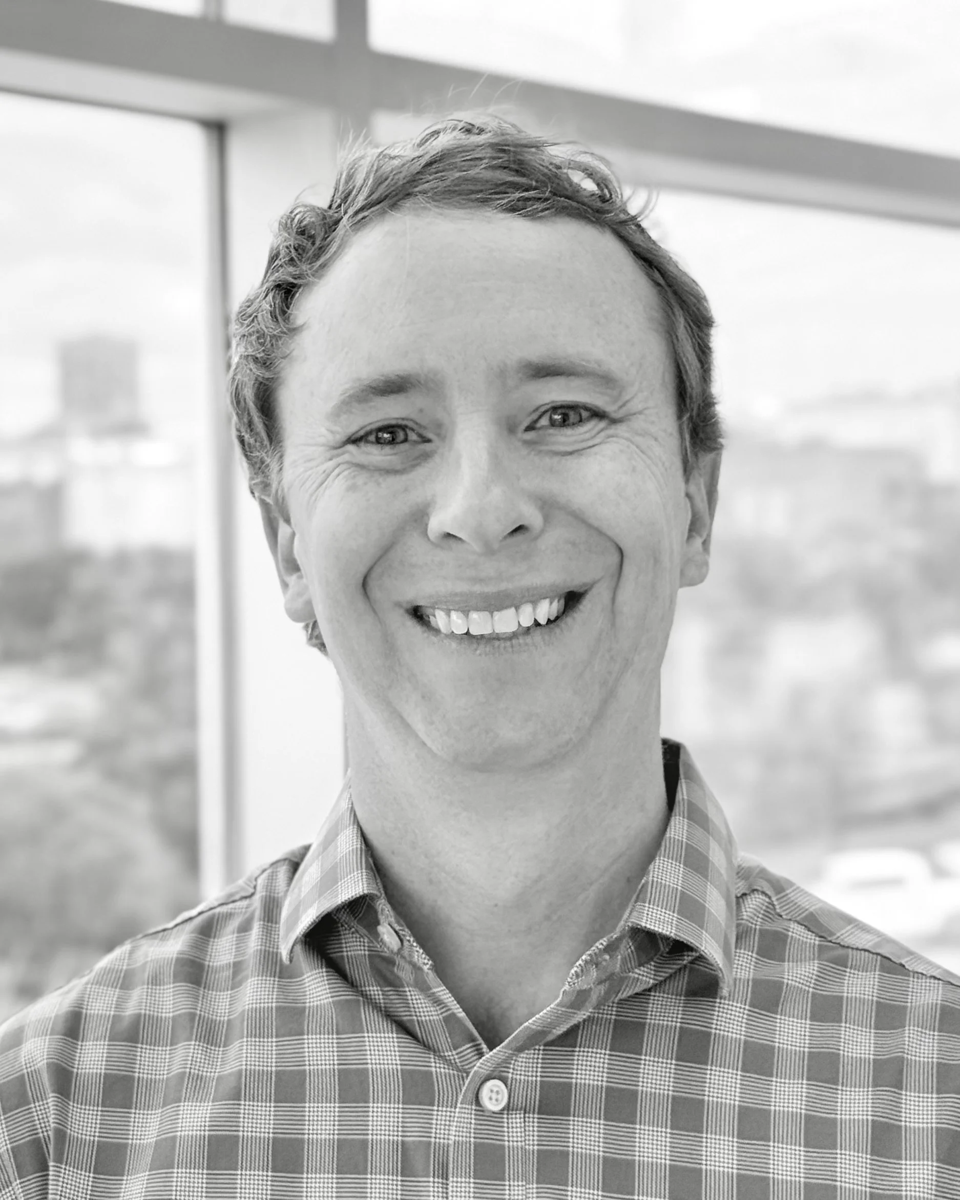 Black and white photo of a smiling man with short wavy hair wearing a checkered shirt, standing indoors in front of large windows with a city skyline in the background.
