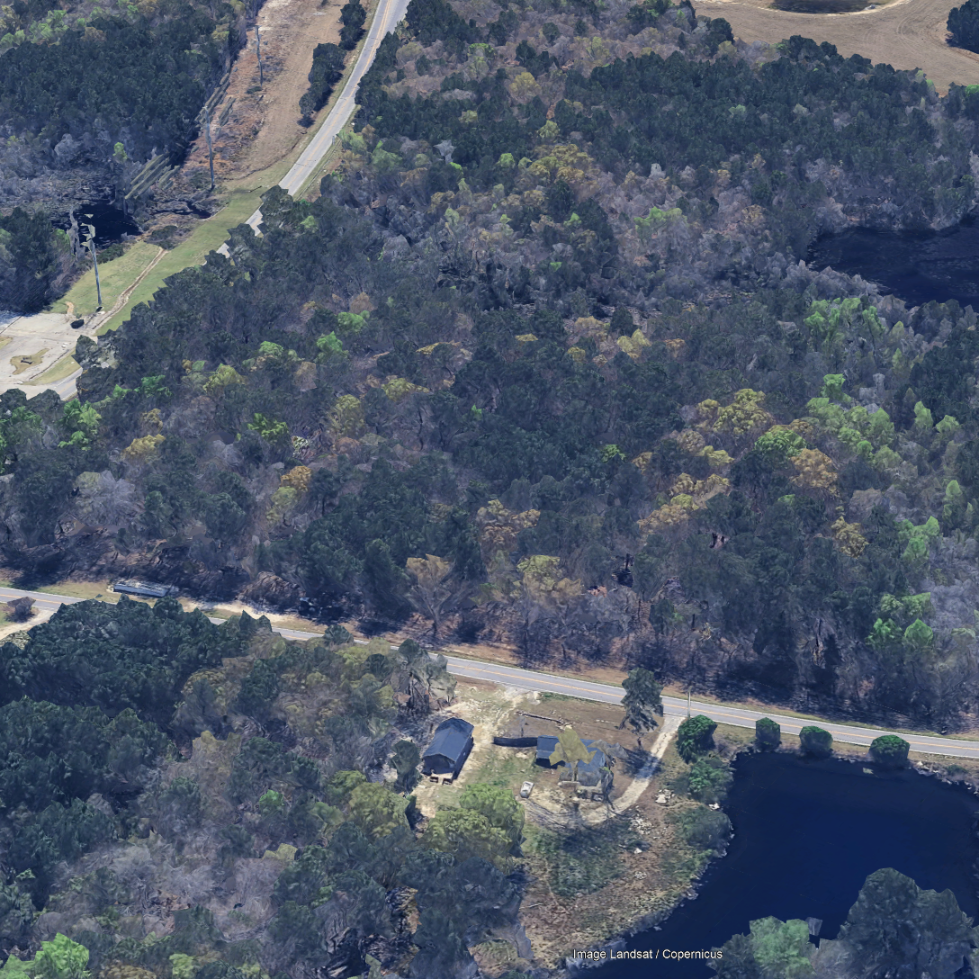 Aerial view of a wooded hillside with a two-lane road cutting through it, small structures near the road, and a lake at the bottom right corner.