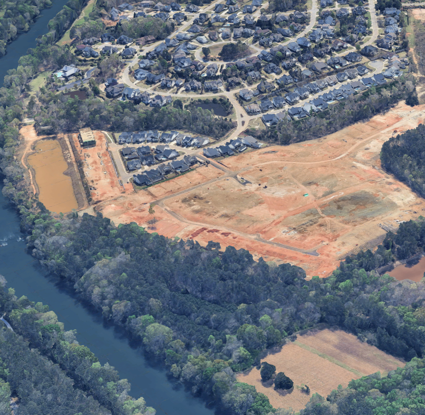 Aerial view of a residential neighborhood with houses, streets, and a construction site with cleared land and dirt roads, adjacent to a river and surrounded by trees.