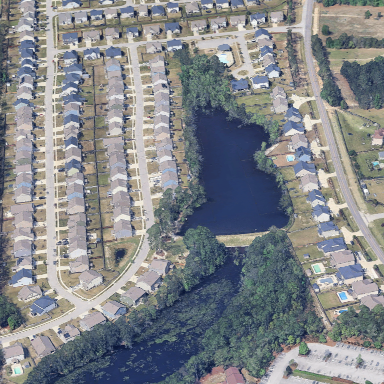 Aerial view of a neighborhood with houses, streets, and a large pond or small lake surrounded by trees.