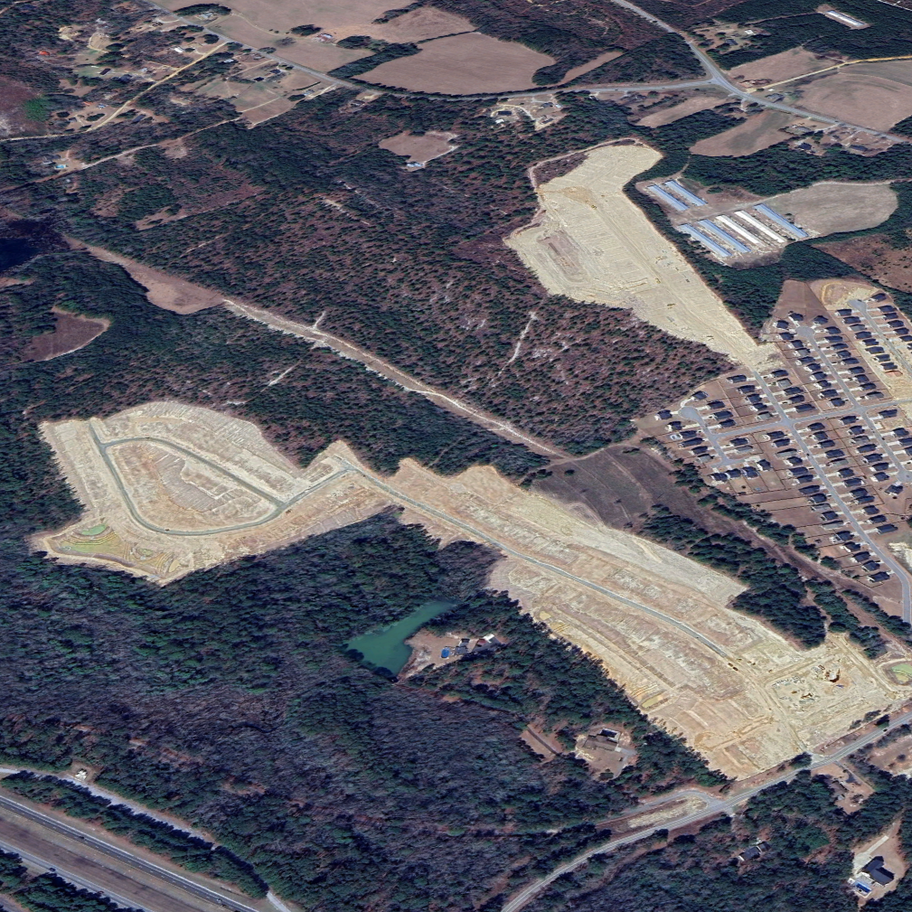 Aerial view of a construction site in a wooded area with partially developed roads and a pond, surrounded by residential neighborhoods and farmland.