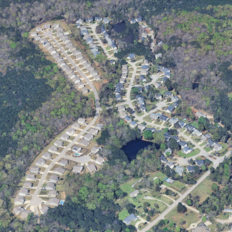 Aerial view of a residential neighborhood with houses, roads, trees, and a small pond.