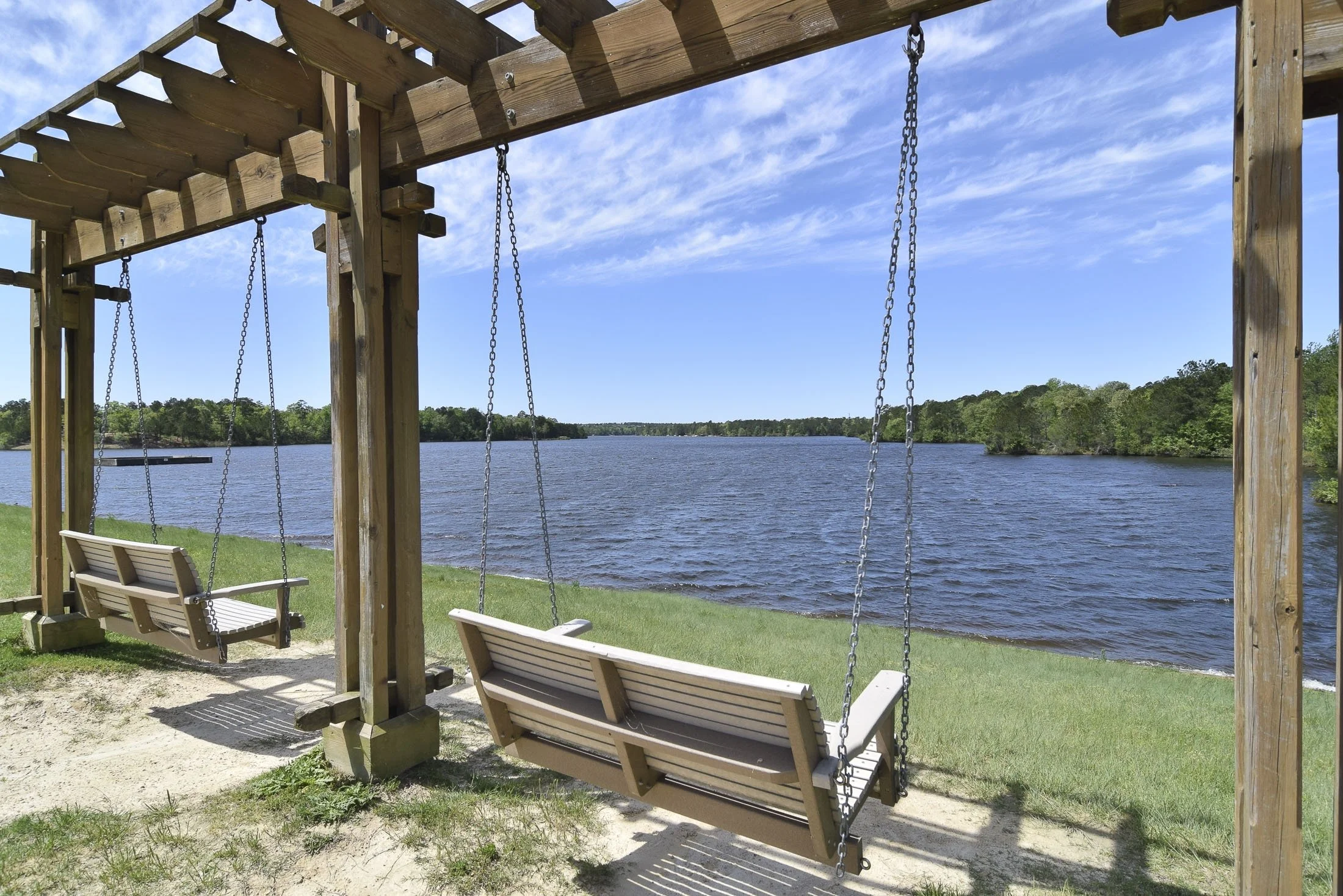 Two wooden swings hanging from a wooden frame overlooking a lake and a grassy shoreline with trees in the distance under a partly cloudy sky.