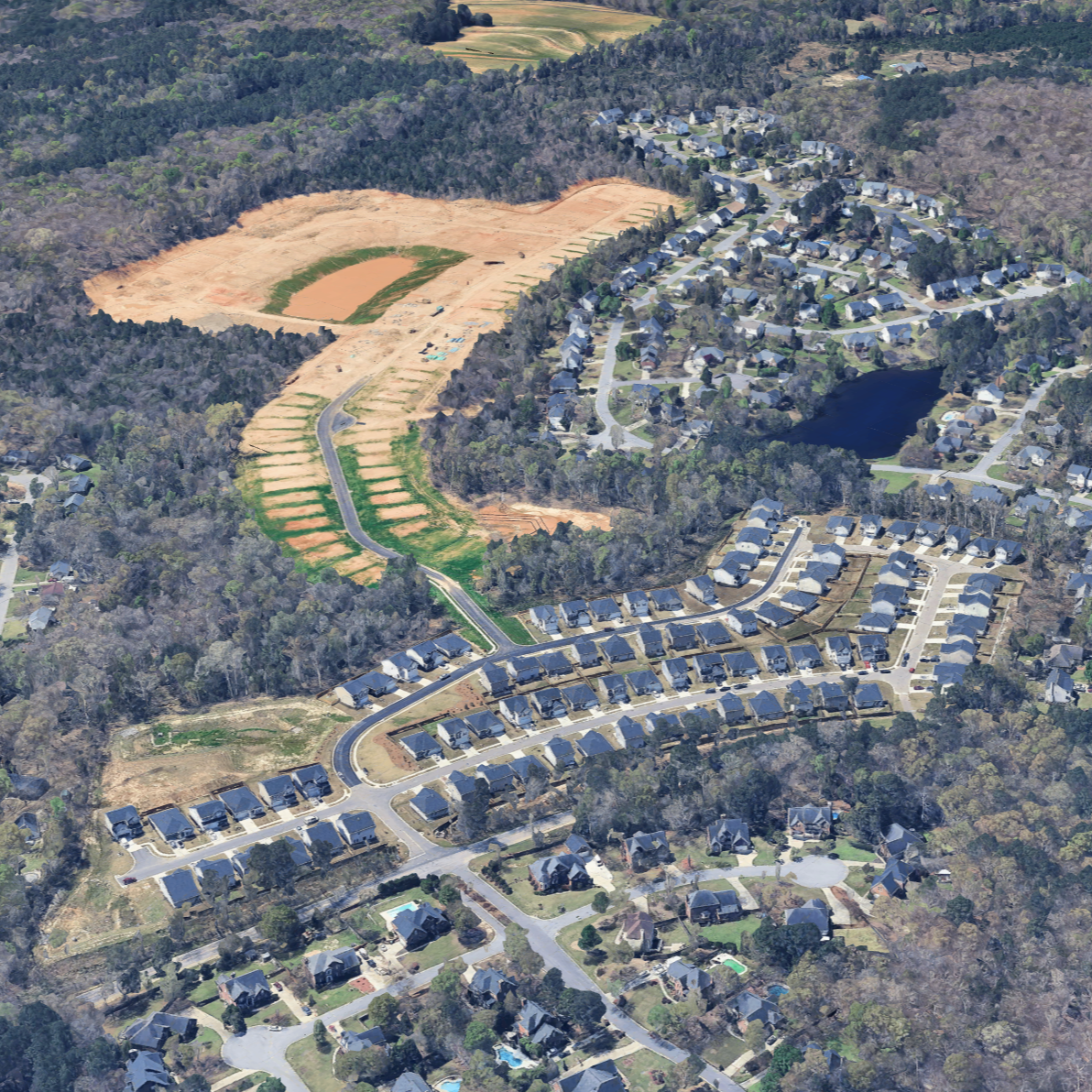 Aerial view of a residential neighborhood with houses and roads surrounded by trees, and a construction site on a hillside with cleared land and pathways.