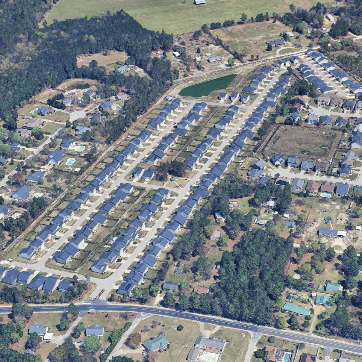 Aerial view of a residential neighborhood with houses, a pond, and green spaces, featuring a mix of single-family homes with yards and swimming pools.