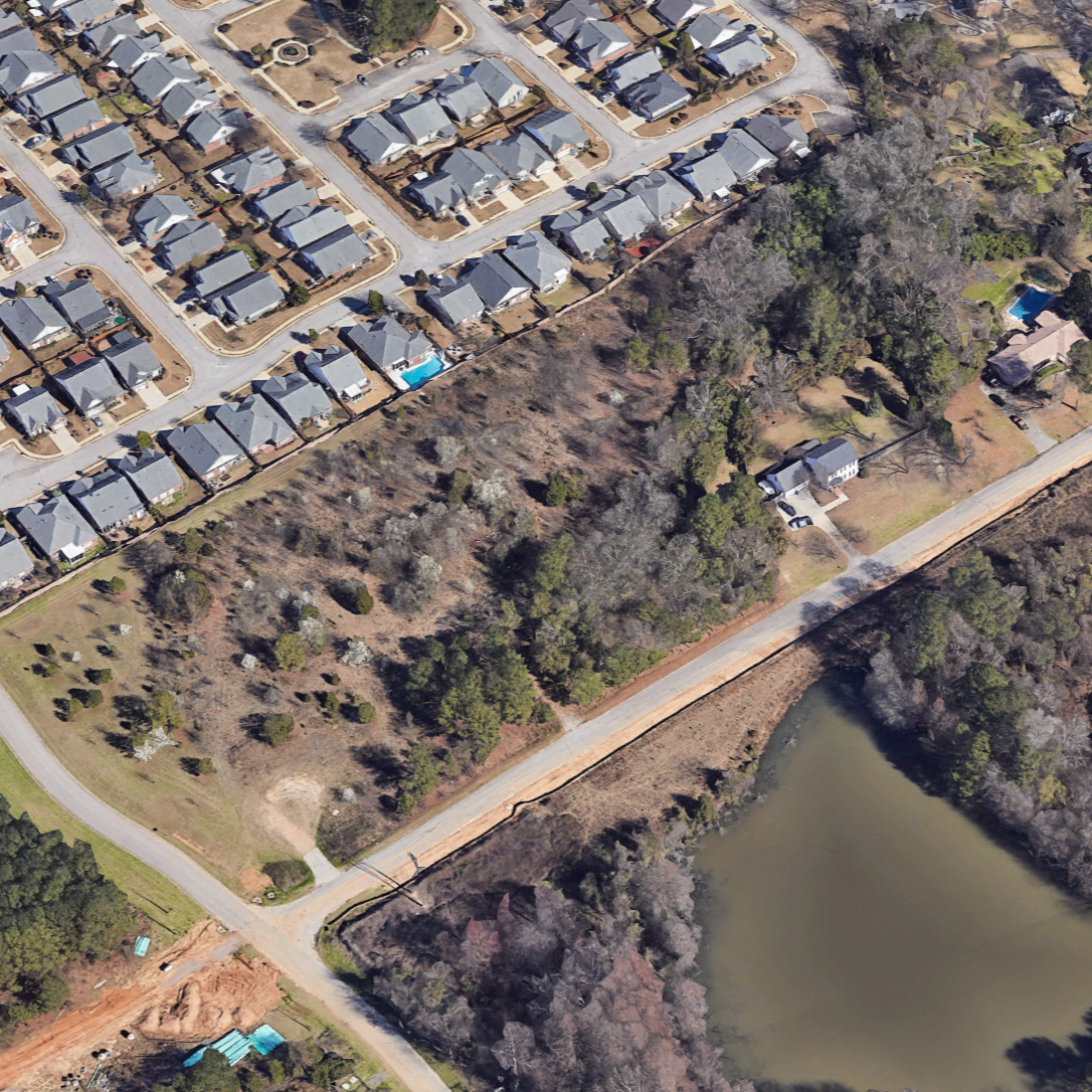 Aerial view of a residential neighborhood with houses, streets, a wooded area, and a pond.