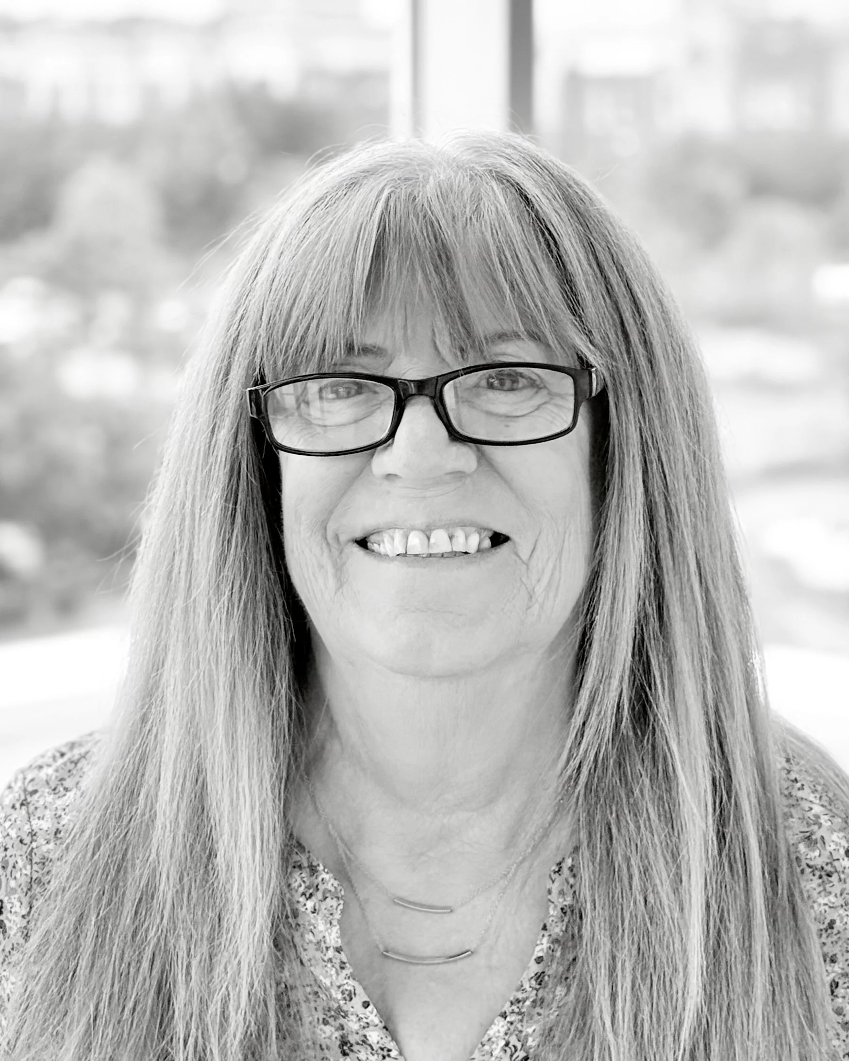 Black and white photo of a smiling woman with long hair, glasses, and layered necklaces, with a blurred outdoor background.