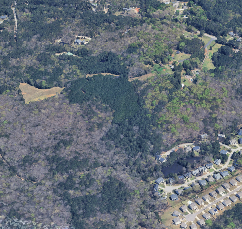 Aerial view of a wooded residential area with houses, tree-covered hills, and some open fields.