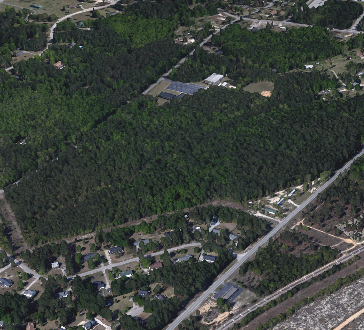 Aerial view of a residential neighborhood with houses, trees, and roads, adjacent to a large wooded area and sports fields.