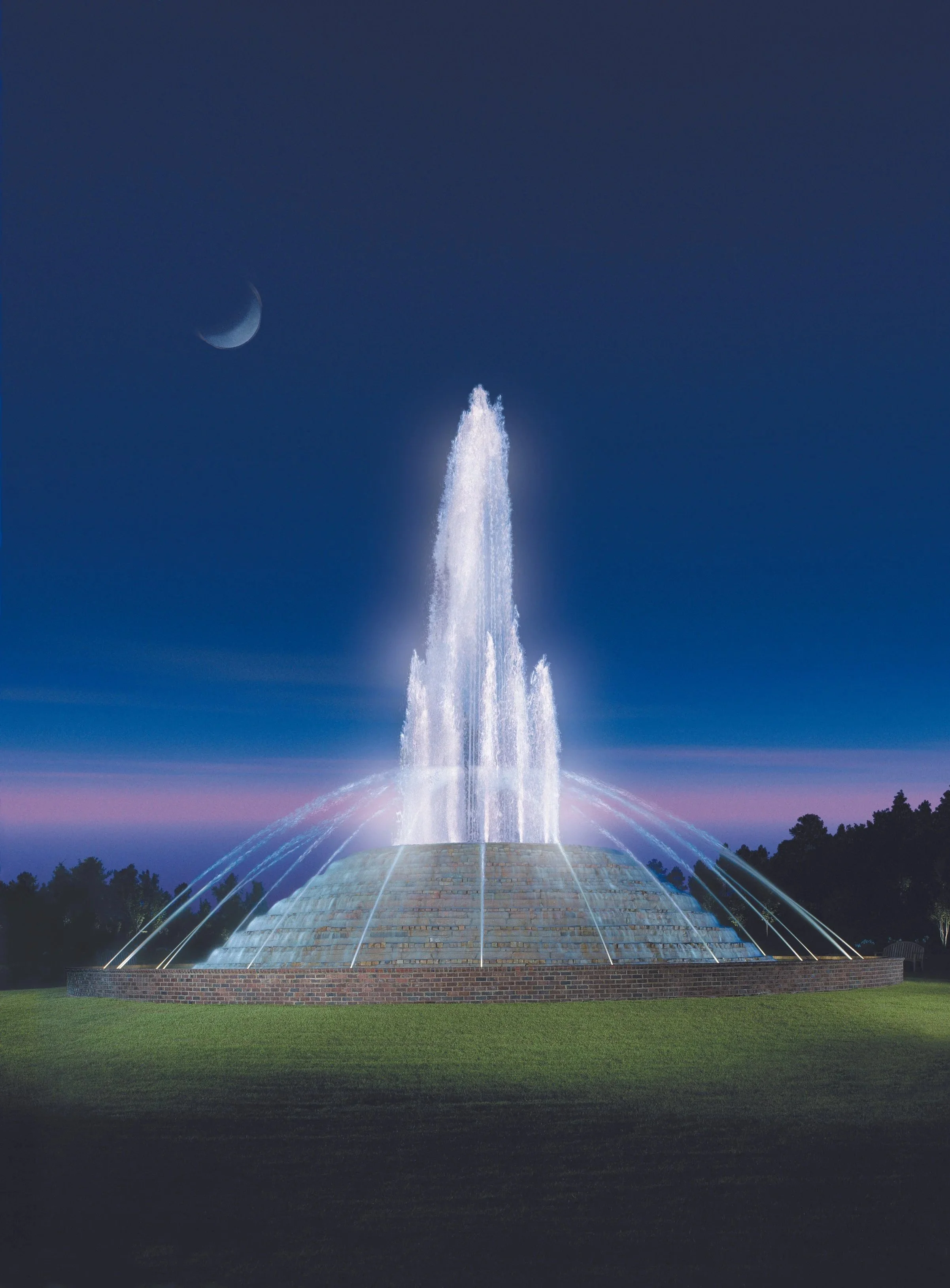A lit fountain at night with water jets shooting upward, modern design, crescent moon in the clear sky, trees in the background, and a grassy area in the foreground.