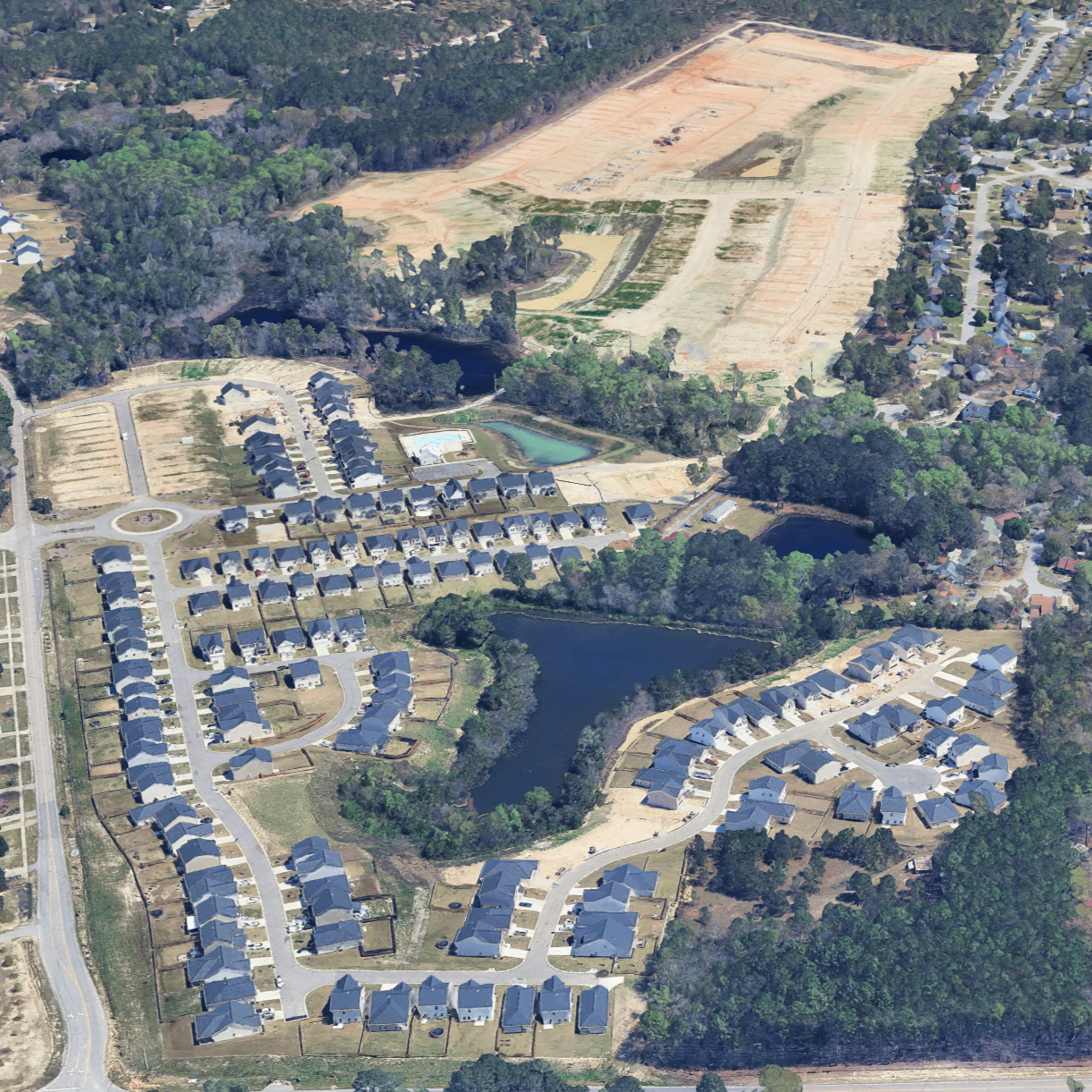 An aerial view of a suburban neighborhood with houses, ponds, and wooded areas alongside a large cleared construction or development site.